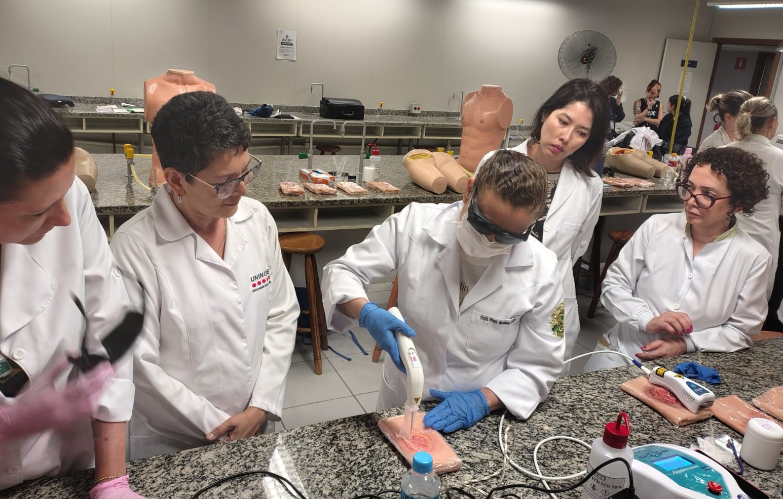 A imagem mostra um grupo de profissionais de saúde, todos vestidos com jalecos brancos, reunidos em uma bancada de laboratório durante uma atividade prática. No centro, uma pessoa utiliza um equipamento portátil — possivelmente um dispositivo de laser terapêutico — aplicado sobre um modelo de pele ou simulador de ferida colocado na bancada. Ela usa óculos de proteção, máscara e luvas azuis, seguindo protocolos de segurança.  As demais pessoas observam atentamente o procedimento, demonstrando interesse e concentração. No ambiente, há diversos materiais de treinamento, incluindo manequins anatômicos, kits e equipamentos distribuídos pelas bancadas ao fundo. A cena sugere uma aula prática ou capacitação voltada para cuidados com feridas ou uso de tecnologias terapêuticas em saúde
