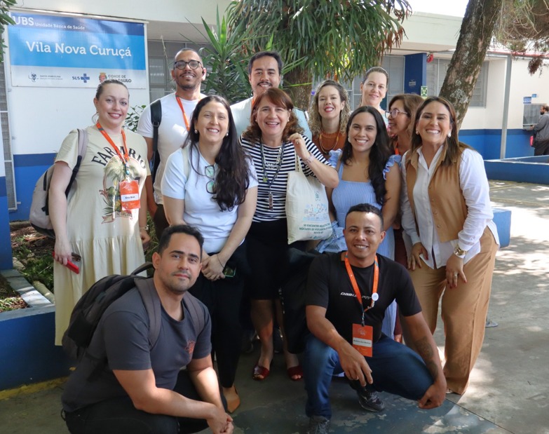A imagem mostra um grupo de cerca de 12 pessoas posando para uma foto em frente a uma unidade de saúde identificada pela placa “UBS Vila Nova Curuçá”. Elas estão reunidas em um ambiente externo, com árvores, plantas e bancos ao redor, sugerindo o pátio da unidade. O grupo é diverso, formado por homens e mulheres, a maioria sorrindo, alguns em pé e outros agachados à frente. Vários usam crachás ou cordões no pescoço, o que indica participação em uma atividade institucional, como visita técnica, capacitação ou intercâmbio profissional. As roupas são casuais ou sociais leves, adequadas a um contexto de trabalho em saúde. Ao fundo, além da placa da UBS, aparecem elementos típicos de um equipamento público de saúde, reforçando o caráter coletivo e profissional do registro.