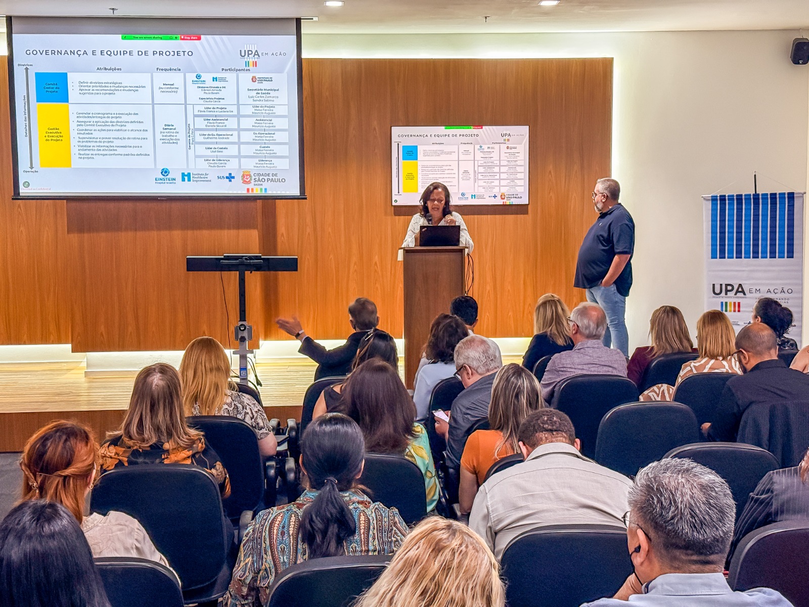 A imagem mostra um auditório durante uma apresentação institucional. Em primeiro plano, há diversas pessoas sentadas em cadeiras, formando a plateia, atentas à atividade. Ao fundo, uma mulher fala ao microfone em um púlpito, utilizando um computador para apoiar a apresentação. Ao lado dela, um homem está em pé, observando. Na parede atrás do palco, dois telões exibem um slide com o título “Governança e equipe de projeto”, relacionado ao programa UPA em Ação, com logotipos institucionais, incluindo o da Prefeitura de São Paulo e de parceiros. O ambiente é formal, típico de um evento técnico ou de prestação de resultados, com iluminação adequada e organização voltada à capacitação ou apresentação de projetos na área da saúde.