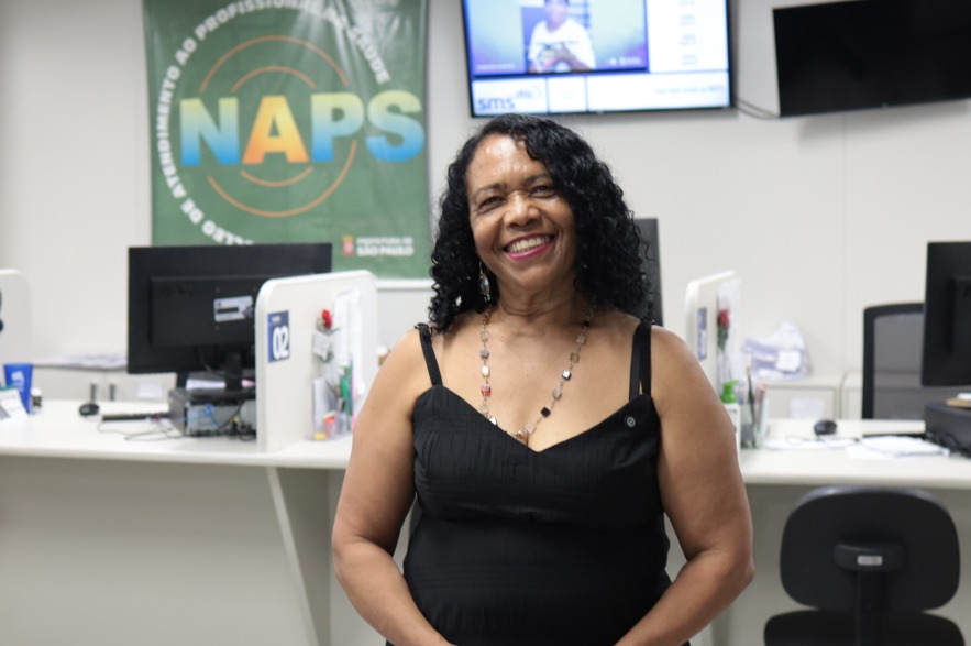 A imagem mostra uma mulher sorridente em primeiro plano, posicionada em um ambiente interno que aparenta ser um serviço de atendimento em saúde. Ela usa um vestido preto sem mangas e um colar delicado, tem cabelos cacheados e soltos, e olha diretamente para a câmera com expressão acolhedora e confiante. Ao fundo, há balcões de atendimento com computadores, cadeiras e materiais de escritório. Em destaque na parede, aparece um banner verde com a sigla NAPS, além de telas de TV fixadas acima dos balcões, exibindo informações institucionais. O espaço é bem iluminado, organizado e remete a um local de acolhimento ou atendimento ao público na área da saúde.