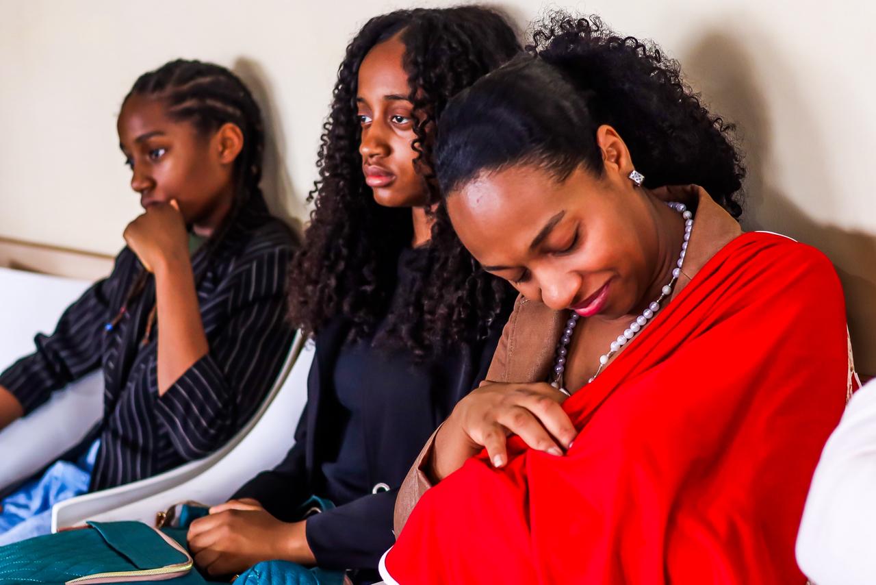 A imagem mostra uma mulher sentada ao lado de três crianças, em um ambiente interno de atendimento. Trata-se de uma mãe acompanhada por três de seus seis filhos, todos vinculados ao acompanhamento realizado pela Casa Angela.  A mulher aparece em primeiro plano, com postura serena e expressão introspectiva, enquanto as crianças estão sentadas próximas, cada uma com gestos e olhares distintos — algumas mais pensativas, outras atentas ao ambiente. A cena transmite um clima de acolhimento, cuidado e vínculo familiar, reforçando a ideia de acompanhamento contínuo e humanizado oferecido pela instituição.