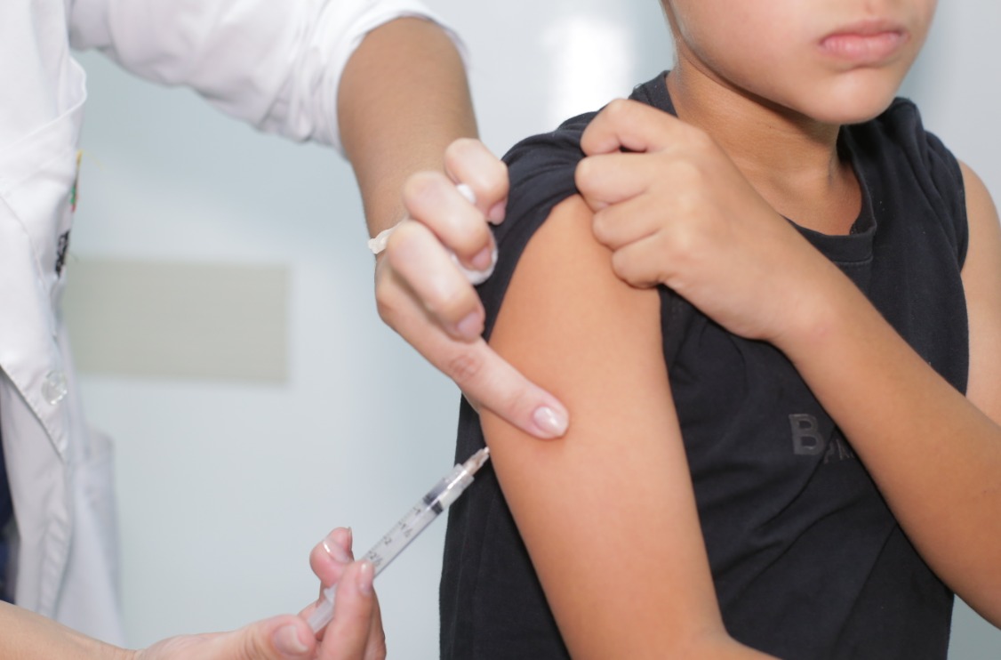 A imagem mostra um menino recebendo uma vacina no braço. Ele veste uma camiseta sem man-gas e segura a manga com a outra mão, deixando o braço exposto. A profissional de saúde, usan-do jaleco branco, segura uma seringa próxima ao braço da criança enquanto aponta o local da aplicação. O ambiente é um local de atendimento médico, com fundo claro e aparência limpa e organizada.