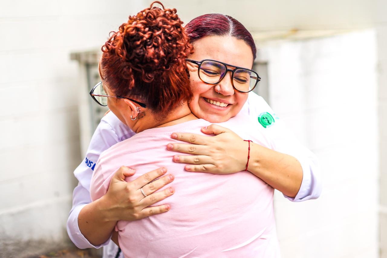 Na imagem, duas mulheres se abraçam em um gesto de acolhimento e afeto. A psicóloga Mariana Ferreira Garcia aparece de frente para a câmera, sorrindo de olhos fechados, transmitindo serenidade e empatia. Ela usa óculos, veste uma blusa clara e mantém os braços envolvidos na outra mulher, num abraço firme e cuidadoso. A participante do grupo Girassóis, grupo de acolhimento às mulheres vítimas de violência doméstica, está de costas para a câmera. Ela veste uma camiseta rosa clara e tem os cabelos presos em um coque. O enquadramento valoriza o gesto do abraço, que ocupa o centro da imagem, reforçando a ideia de cuidado, escuta e apoio emocional. O fundo é neutro e claro, sem muitos elementos, o que direciona o olhar para o encontro entre as duas mulheres e para a potência simbólica do acolhimento oferecido pelo grupo.