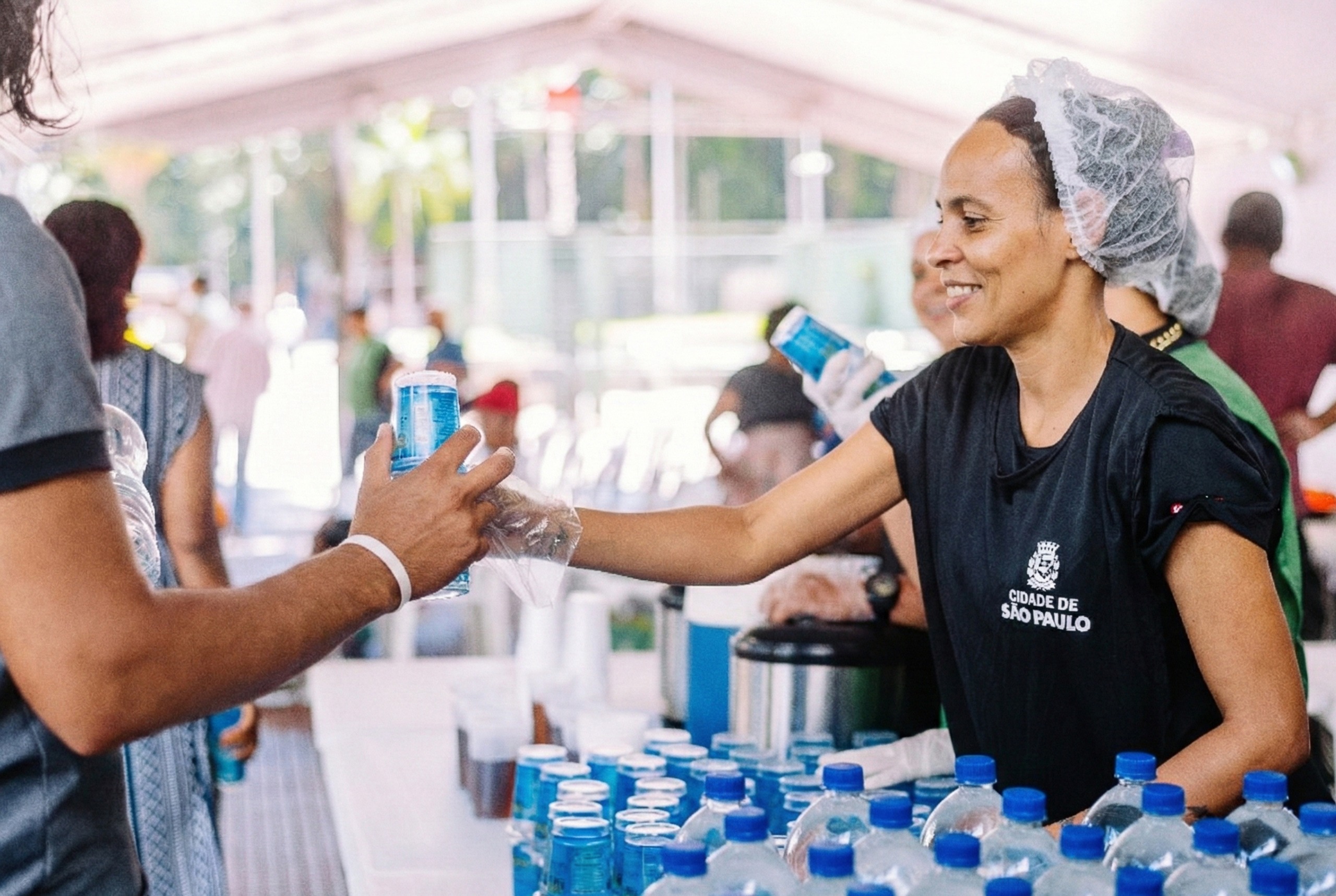 A imagem mostra uma cena de atendimento ao público sob uma tenda da Operação Altas Temperaturas. Em primeiro plano, uma mulher sorri enquanto entrega um copo plástico com água a outra pessoa. Ela usa touca descartável, luvas transparentes e uma camiseta preta com o brasão e a inscrição “Cidade de São Paulo”. Sobre a mesa à sua frente há várias garrafas de água e latas organizadas, além de copos plásticos