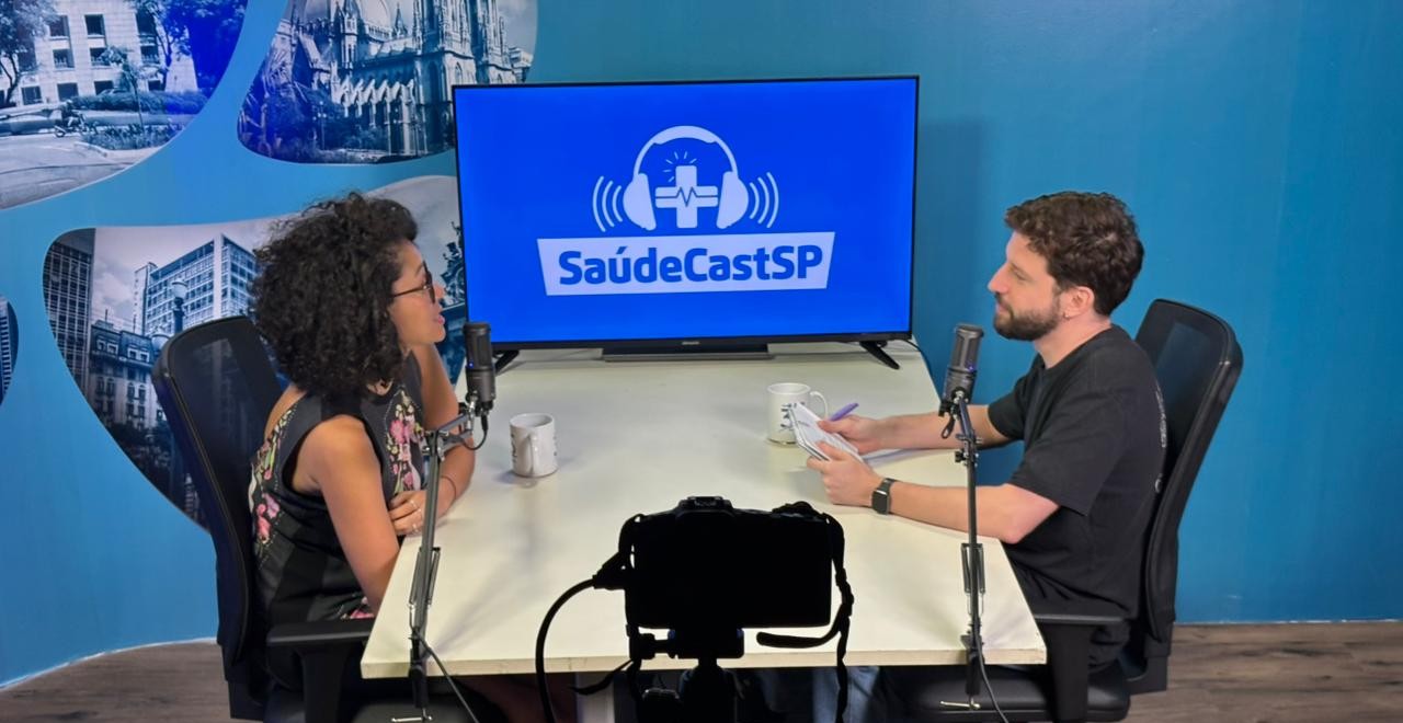 A imagem mostra a gravação de um podcast em um estúdio com paredes azuis e detalhes que remetem à cidade de São Paulo. Dois participantes, uma mulher com cabelos cacheados à esquerda e um homem à direita, estão sentados frente a frente em uma mesa branca, equipada com microfones profissionais e canecas. Ao fundo, uma tela de televisão exibe o logotipo 