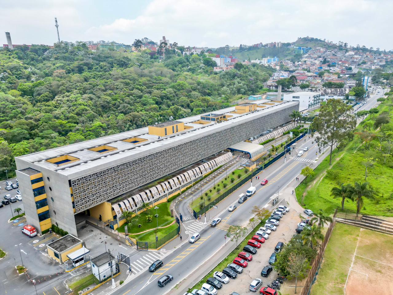 Vista panorâmica do Hospital Municipal Cidade Tiradentes (Hospital Municipal Carmen Prudente), destacando sua arquitetura linear e sua inserção no relevo da Zona Leste de São Paulo. O hospital está situado ao pé de uma encosta densamente arborizada, criando um contraste entre a construção cinza e o verde da vegetação nativa ao fundo. Ao fundo e nas laterais, observa-se o tecido urbano característico da região, com casas e prédios baixos distribuídos pelas colinas da Cidade Tiradentes.