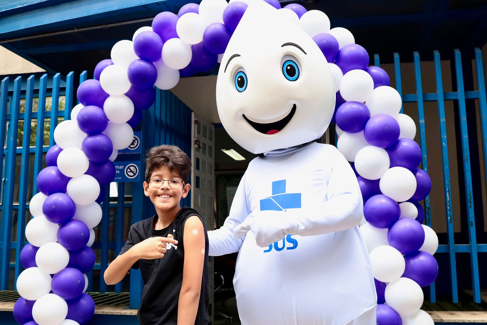 A imagem mostra uma criança sorridente usando óculos, em frente à entrada de uma Unidade Básica de Saúde (UBS) decorada com um arco de balões roxos e brancos. Ela aponta para o próprio braço, onde dá para ver curativos de vacinação. Ao lado da criança há uma pessoa usando uma fantasia de Zé Gotinha, o mascote da vacinação que tem cabeça em formato arredondado, olhos azuis grandes e um sorriso bem amigável. O mascote também aponta para o braço da criança, que recebeu a vacina. 