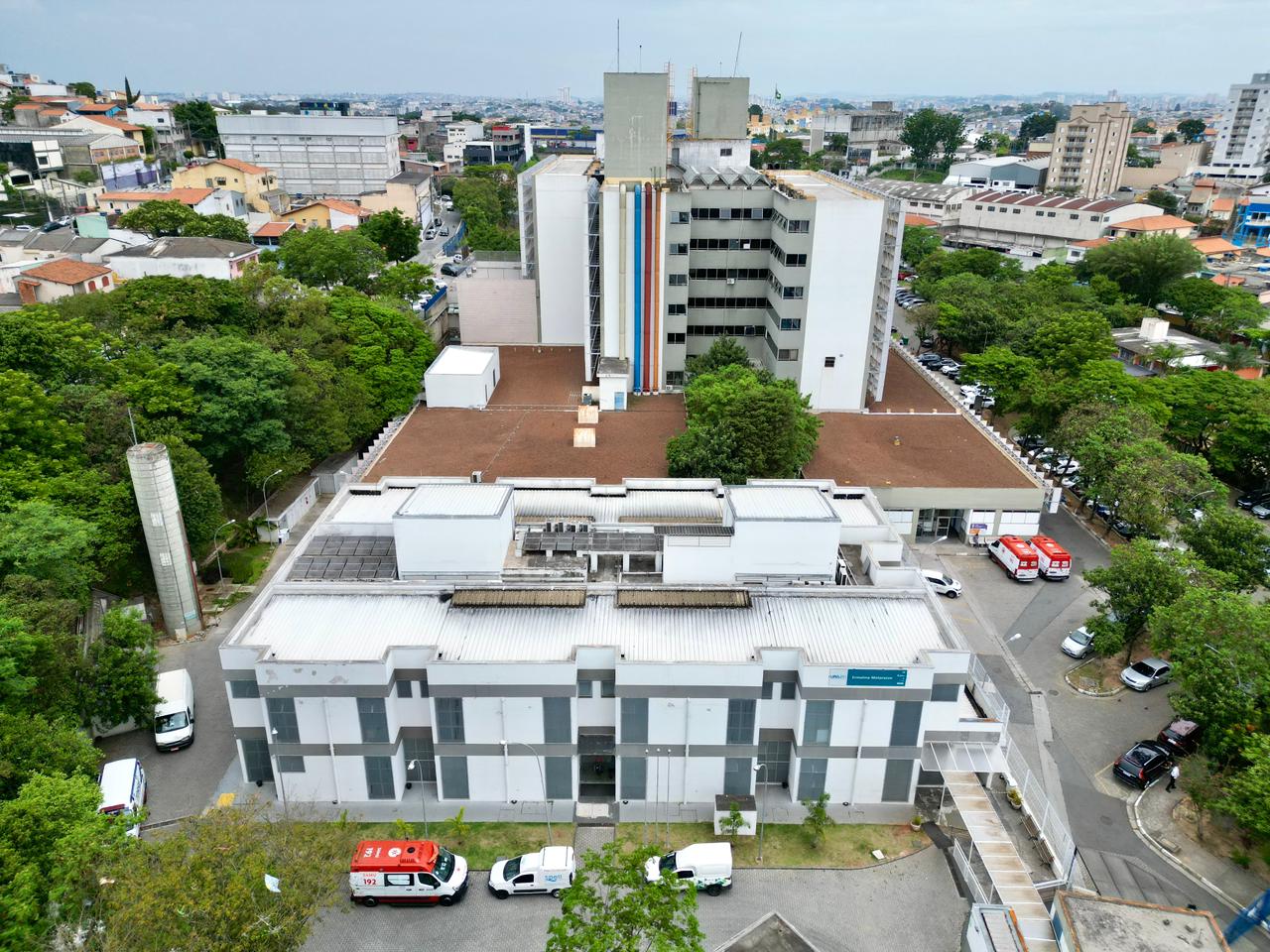 Foto aérea, feita por drone, da Unidade de Pronto Atendimento (UPA) Ermelino Matarazzo. O prédio principal é alto, com fachada clara e listras verticais coloridas no centro. Outros blocos anexos mais baixos, com telhados planos e aparência funcional. Ambulâncias estão estacionadas na parte inferior direita e também na frente.