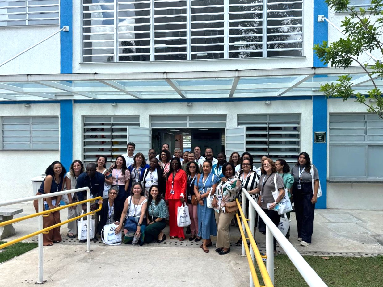 A imagem mostra um grupo numeroso de pessoas posando para uma foto coletiva em frente a um prédio institucional, possivelmente uma unidade de saúde ou centro público. O edifício tem fachada branca com detalhes em azul, janelas amplas com venezianas metálicas e uma marquise de vidro sobre a entrada principal. O grupo está reunido na área externa, em frente à porta aberta, sobre uma rampa de acesso com corrimãos amarelos, o que indica preocupação com acessibilidade. As pessoas estão bem próximas umas das outras, sorrindo, em clima de confraternização ou encerramento de atividade. Há diversidade de gênero, idade e etnia, e muitos usam crachás no pescoço, sugerindo que participam de um evento institucional, capacitação, visita técnica ou encontro profissional. Algumas seguram sacolas personalizadas, o que reforça a ideia de um evento organizado. O vestuário é variado, indo do casual ao social, com destaque para uma pessoa ao centro usando roupa vermelha, que chama atenção visualmente. O ambiente é diurno, com luz natural, árvores ao redor e aparência organizada e acolhedora. No conjunto, a imagem transmite integração, trabalho em equipe e caráter institucional, possivelmente relacionada a uma ação de saúde, formação ou cooperação profissional.