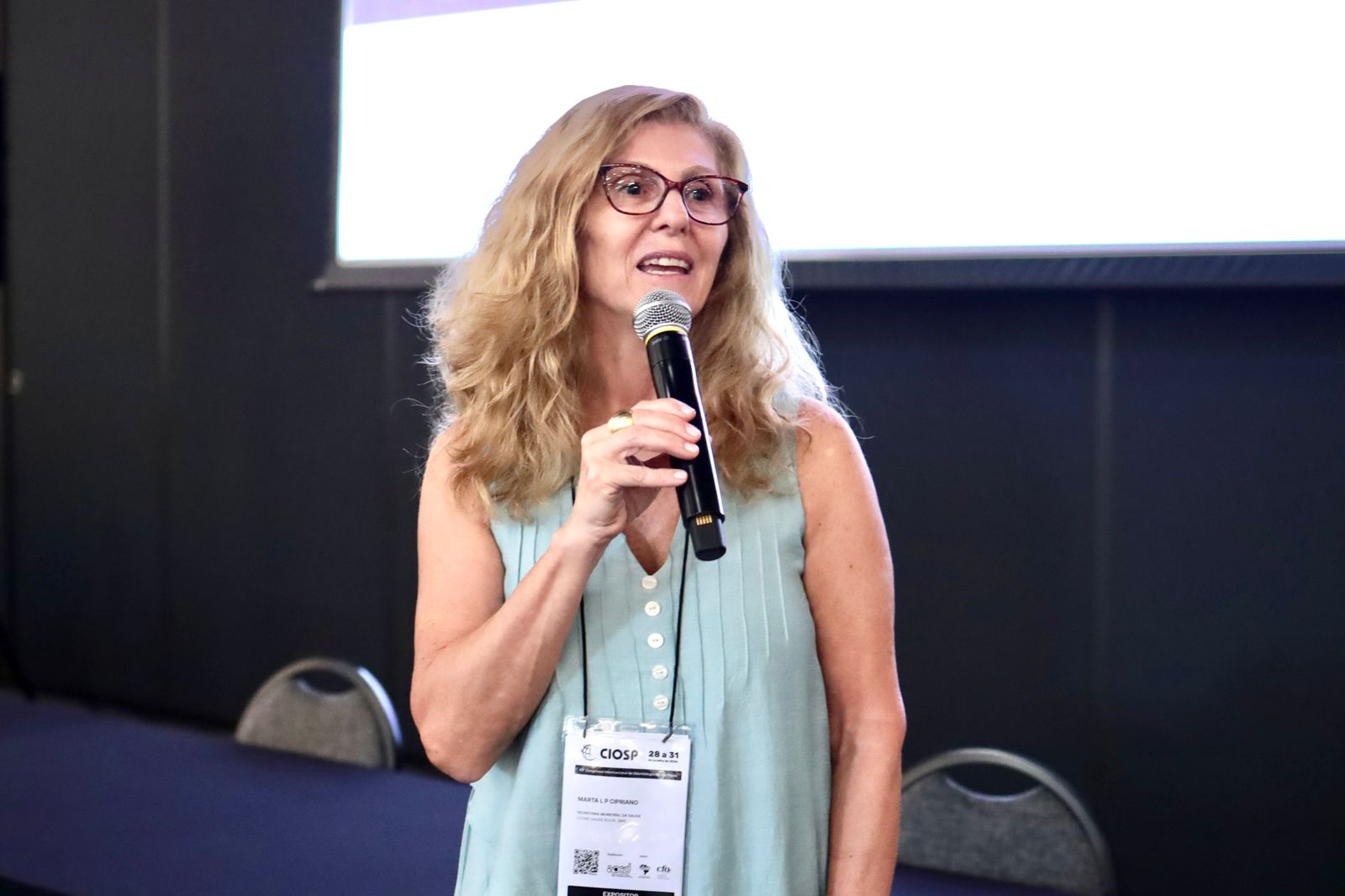 A imagem mostra uma mulher falando ao microfone, em um ambiente de evento ou congresso. Ela está em pé, com postura confiante, segurando um microfone sem fio, como se estivesse fazendo uma palestra, apresentação ou fala pública.  A mulher tem cabelos loiros, ondulados, na altura dos ombros, usa óculos de armação escura e veste uma blusa sem mangas em tom claro esverdeado. No pescoço, há um crachá de identificação, típico de eventos profissionais, indicando que ela participa oficialmente da atividade, possivelmente como palestrante ou mediadora.  Ao fundo, vê-se uma mesa com cadeiras e uma tela de projeção, reforçando o contexto de auditório ou sala de conferências. A expressão facial sugere que ela está engajada e concentrada, falando para um público fora do enquadramento.