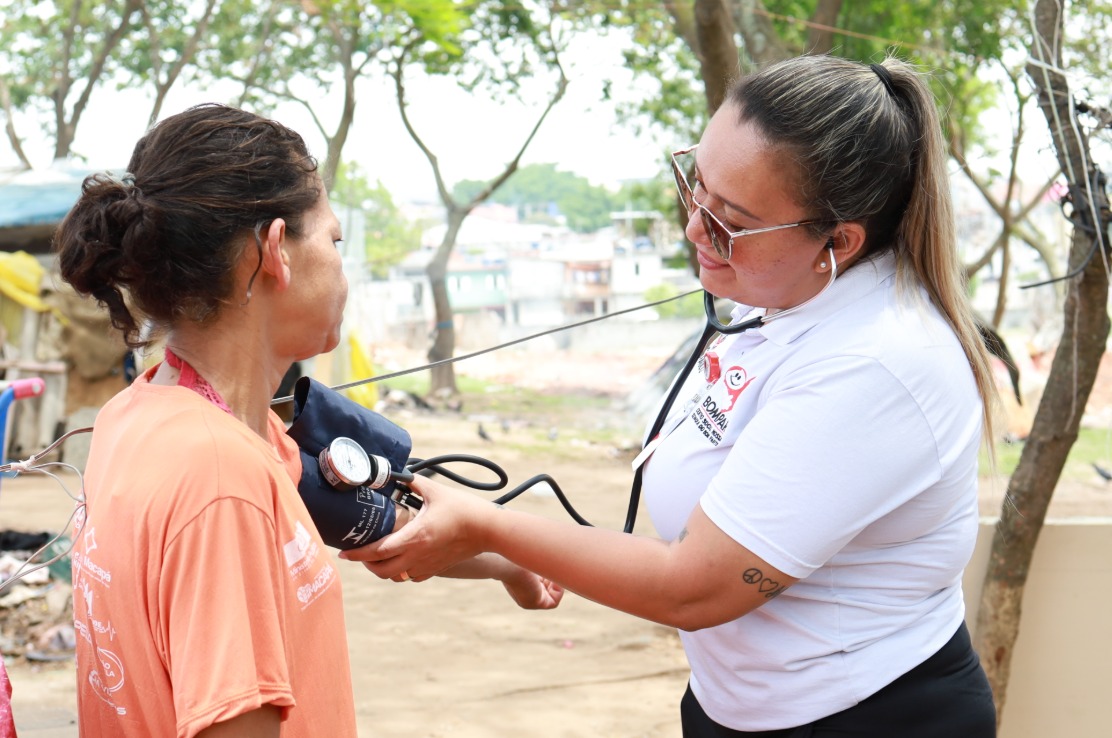  A imagem mostra uma profissional de saúde realizando a aferição da pressão arterial de uma mulher em um ambiente ao ar livre. A profissional, que veste uma camisa branca com logotipos de um projeto de assistência, utiliza um estetoscópio e um esfigmomanômetro (aparelho de pressão) para examinar a paciente, que está vestida com uma camiseta laranja. A cena sugere um atendimento comunitário ou ação social de saúde, possivelmente em uma área de vulnerabilidade. Ao fundo, há árvores, construções e sinais de uma ocupação urbana informal. A interação transmite cuidado, empatia e atenção às necessidades básicas de saúde da população