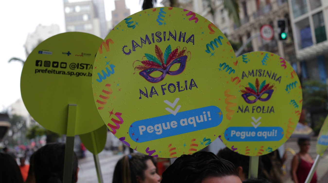 A imagem mostra pessoas em um bloco de Carnaval, segurando placas circulares verdes com a mensagem “Camisinha na Folia – Pegue aqui!”.  No centro das placas há a ilustração de uma máscara carnavalesca colorida, com penas e elementos festivos, reforçando o clima de festa. O design inclui confetes e serpentinas em tons vibrantes, associados à celebração.  Ao fundo, aparecem prédios, semáforos e parte da multidão. Em uma das placas, é possível ver o endereço prefeitura.sp.gov.br, pois se trata de uma campanha institucional da Prefeitura de São Paulo.