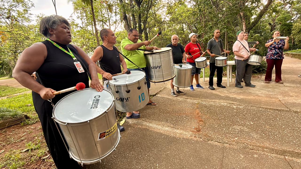 A imagem mostra o bloco de Carnaval Bibitantã tocando instrumentos em um parque. Cercados por árvores, cada pessoa segura um instrumento. Os instrumentos são grandes e prateados. À esquerda da imagem, uma mulher veste roupa preta e segura uma baqueta com ponta vermelha, posicionada sobre o tambor. Ao lado dela, há outros homens e mulheres, a maioria usando roupas casuais como camisetas, regatas e bonés. Um dos homens, no centro, veste camiseta verde e toca caixa.