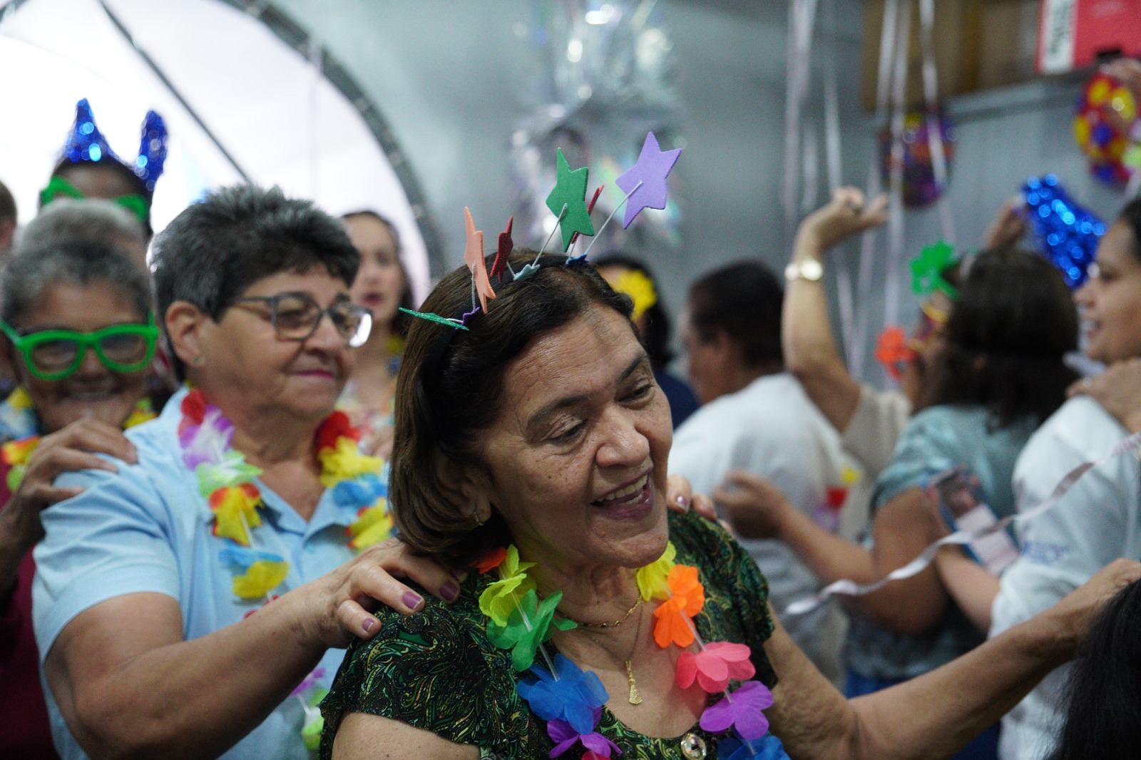 A imagem mostra um grupo de mulheres, em sua maioria idosas, participando de uma atividade festiva em ambiente fechado, provavelmente em um evento comunitário ou institucional. No centro da cena, uma mulher sorri enquanto dança, usando um adereço colorido na cabeça em formato de estrelas e um colar havaiano de flores artificiais. Ela veste uma blusa verde estampada e aparenta estar se divertindo com o momento. Atrás dela, outra mulher, de camisa azul-clara, coloca as mãos em seus ombros, formando uma fila, como em uma dança em grupo. Ela também usa acessórios festivos, como óculos verdes e um colar colorido. Outras participantes aparecem ao fundo, com enfeites brilhantes, chapéus e serpentinas, reforçando o clima de comemoração. O ambiente é decorado com fitas, balões e elementos cintilantes, criando uma atmosfera alegre e descontraída. As expressões das pessoas indicam animação, integração e bem-estar.