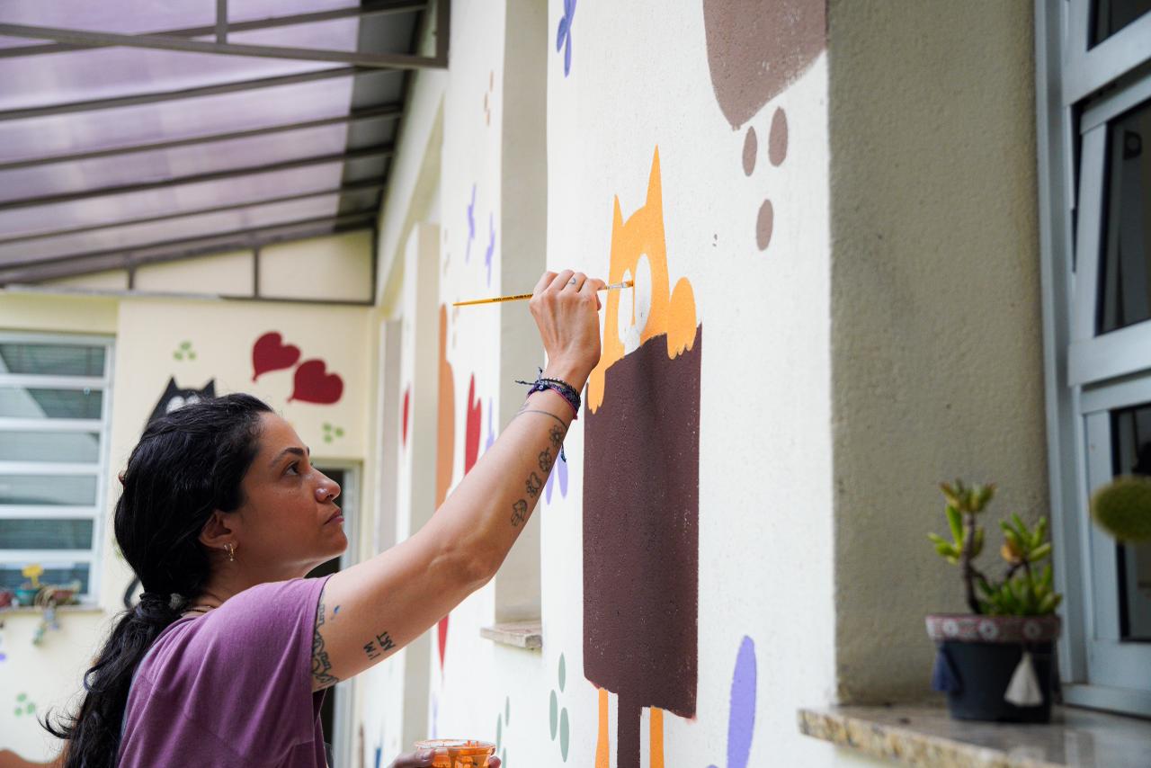 A imagem mostra uma mulher pintando um mural em uma parede externa, possivelmente em um ambiente escolar ou comunitário. Ela segura um pincel com a mão direita e aplica tinta em um desenho colorido, que parece representar personagens ou formas lúdicas. Na outra mão, segura um pequeno recipiente com tinta. A mulher veste uma camiseta roxa, tem tatuagens nos braços e o cabelo preso. Sua expressão é concentrada, indicando atenção aos detalhes do trabalho. Ao fundo, há janelas, vasos de plantas e outras pinturas na parede, com corações e figuras decorativas, reforçando o caráter artístico e acolhedor do espaço.