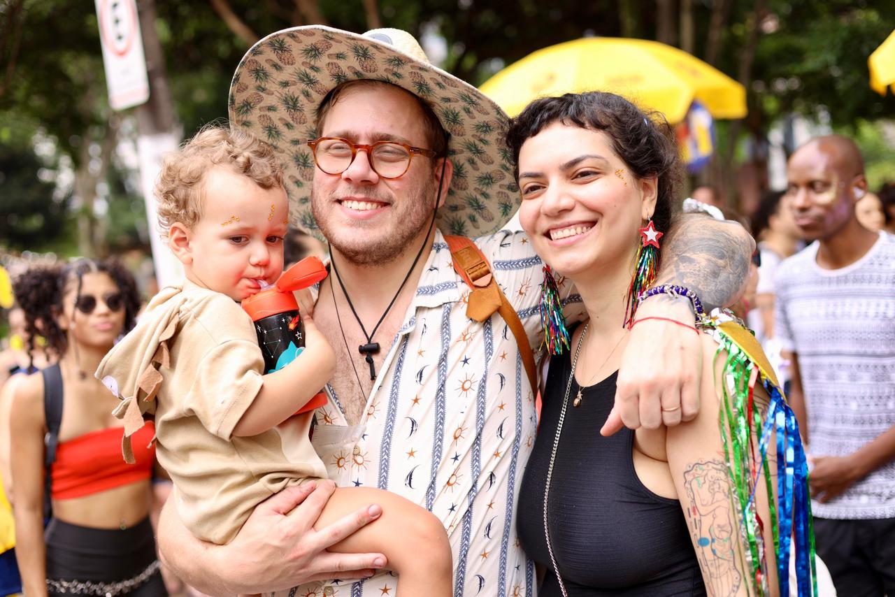 A imagem mostra três pessoas em primeiro plano, em meio a um ambiente festivo ao ar livre, que parece ser um bloco de Carnaval ou uma celebração popular. No centro, há um homem sorrindo, usando óculos, chapéu estampado e camisa clara com desenhos. Ele segura no colo uma criança pequena, que parece estar bebendo água de uma garrafinha vermelha. A criança veste uma roupa bege e tem glitter no rosto, típico de festas carnavalescas. À direita, há uma mulher sorridente, com o braço em volta do homem. Ela usa uma blusa preta sem mangas, brincos coloridos em forma de estrela e tem tatuagens visíveis no braço. Também carrega enfeites coloridos pendurados no ombro, reforçando o clima festivo. Ao fundo, aparecem outras pessoas participando do evento, além de árvores e guarda-sóis amarelos,