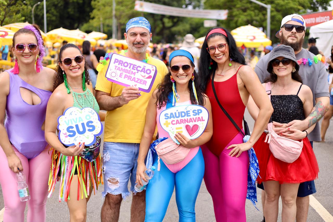 A imagem mostra um grupo de oito pessoas posando juntas em meio a um evento festivo ao ar livre, provavelmente um bloco de Carnaval. Todas estão sorrindo e voltadas para a câmera, transmitindo um clima de alegria, descontração e integração. As roupas são bastante coloridas e chamativas, com predominância de collants, peças justas, brilhos, franjas e acessórios típicos do Carnaval. No centro, um homem segura uma placa com a frase “Proteção também é prazer”, acompanhada de um desenho de preservativo, reforçando uma mensagem de prevenção e cuidado com a saúde. Duas mulheres também seguram placas: uma com “Viva o SUS” e outra com “Saúde SP + Carnaval”, com um coração, destacando a valorização do sistema público de saúde e ações de conscientização durante a festa.
