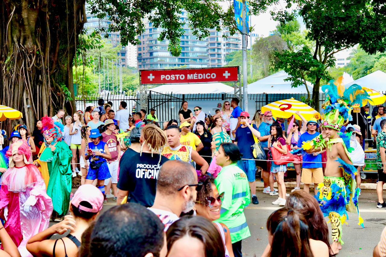 A imagem retrata um carnaval de rua em São Paulo. Há uma grande concentração de pessoas na rua, muitas usando fantasias coloridas, com penas, brilhos e adereços típicos do carnaval. O clima é festivo e descontraído, com foliões de diferentes idades aproveitando a celebração ao ar livre. Ao fundo, aparece uma estrutura com a placa “Posto Médico”, comum em grandes blocos de carnaval para atendimento emergencial. Também é possível ver prédios altos e bastante arborização, características de algumas regiões da cidade. A cena transmite a energia vibrante dos blocos paulistanos, com mistura de fantasia, música e multidão ocupando as ruas.