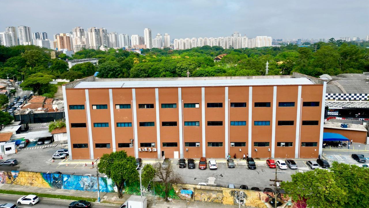 A imagem mostra uma vista aérea de um grande edifício retangular, de vários andares, com fachada em tons de marrom e faixas verticais claras. Em frente ao prédio, há uma área de estacionamento com diversos carros alinhados. Na parte inferior da imagem, vê-se um muro grafitado com desenhos coloridos e uma rua com circulação de veículos. Ao fundo, há uma extensa área verde, com muitas árvores, que separa o edifício de um conjunto de prédios residenciais altos, formando o horizonte urbano. O céu está claro, indicando um dia ensolarado.