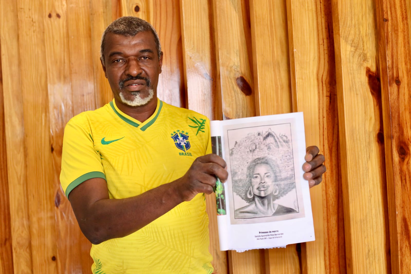 A imagem mostra um homem de meia-idade em pé diante de uma parede de madeira clara, composta por tábuas verticais. Ele veste uma camisa amarela da seleção brasileira de futebol, com detalhes em verde. O homem segura com as duas mãos um desenho em preto e branco de uma mulher com cabelo volumoso estilo afro, usando brincos grandes e com expressão serena. A arte parece ser um retrato detalhado, feito a lápis.