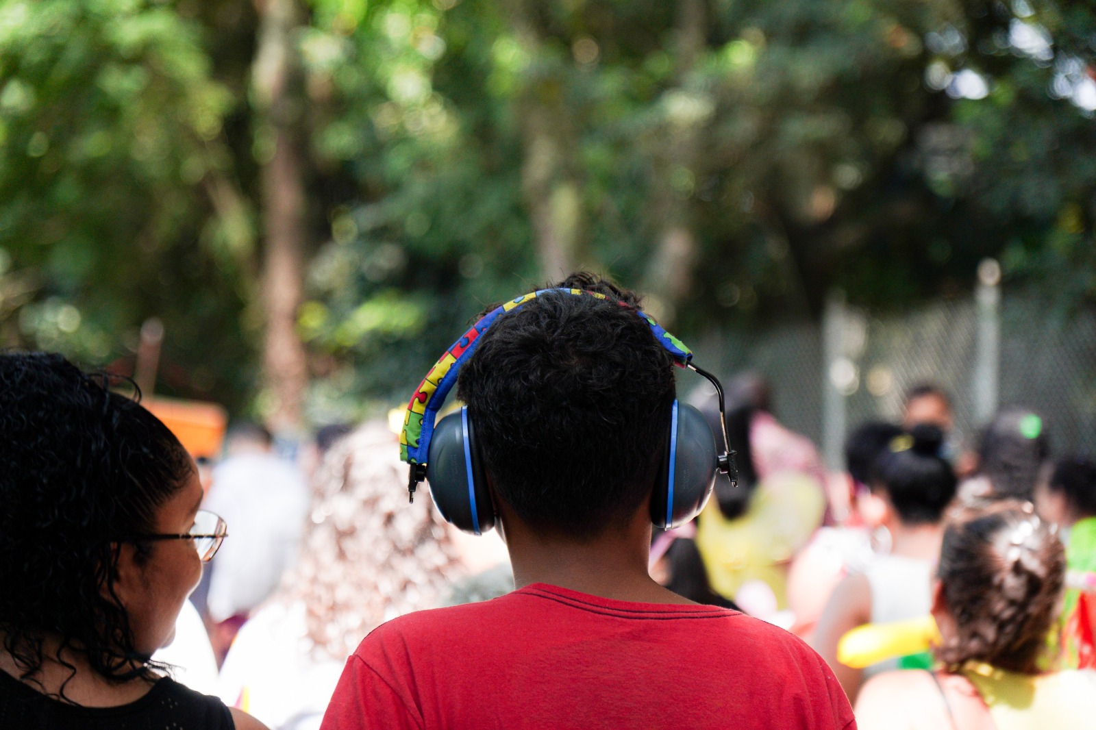 A imagem mostra, em primeiro plano, uma criança vista de costas, usando uma camiseta vermelha e protetores auriculares grandes e coloridos, que cobrem totalmente os ouvidos. Ao lado dela, aparece parcialmente uma mulher, também vista de perfil. Ao fundo, desfocado, há um grupo de pessoas reunidas ao ar livre, em um ambiente arborizado, sugerindo um evento ou atividade em um parque ou espaço público. A cena transmite a ideia de um momento coletivo, possivelmente festivo ou cultural, com atenção especial ao cuidado com o conforto sensorial da criança.