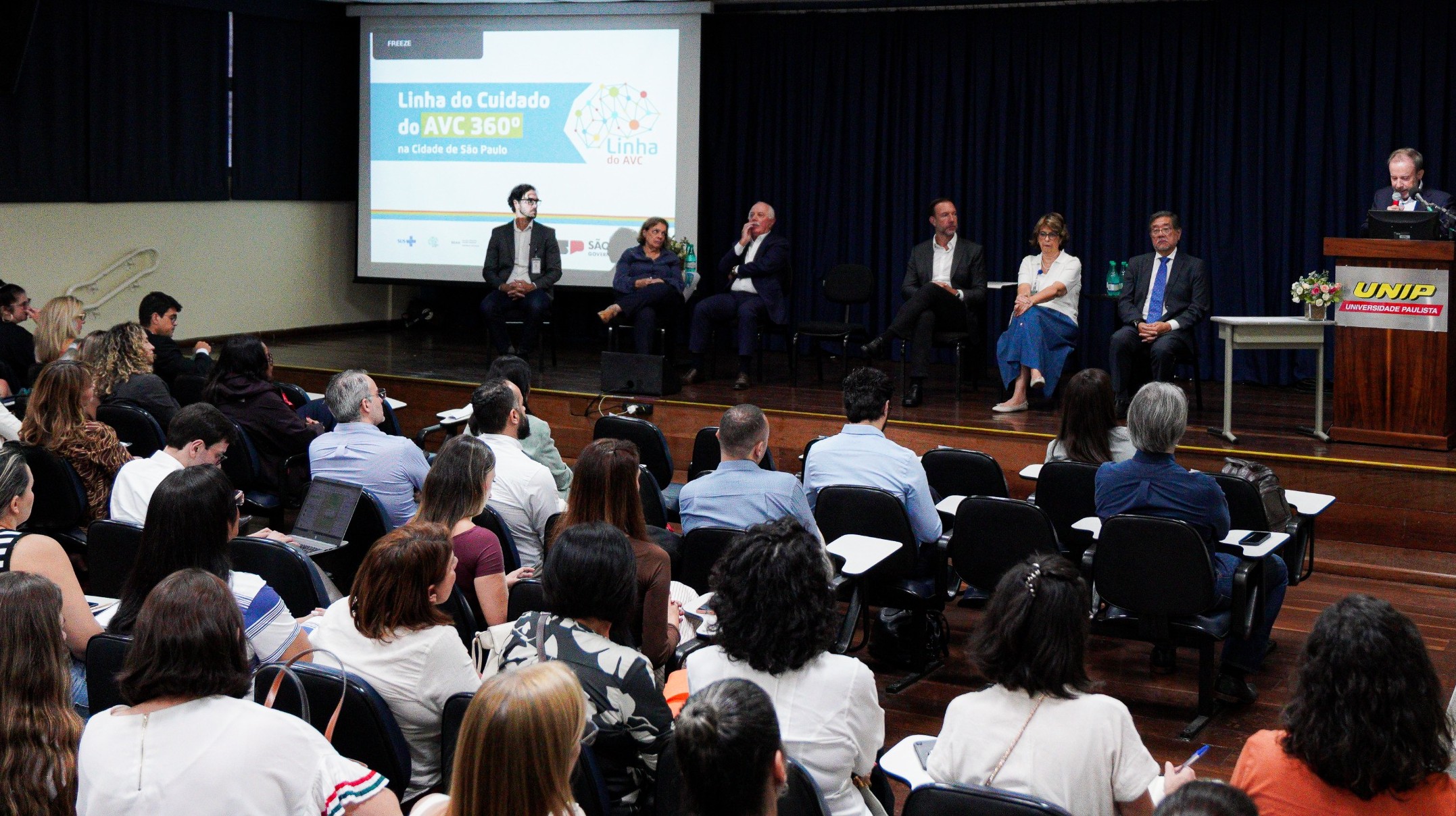A imagem mostra um auditório lotado durante um evento ou seminário na área da saúde. No palco, há uma mesa com vários especialistas sentados, enquanto um homem fala ao púlpito, identificado com o logotipo da UNIP (Universidade Paulista). Ao fundo, um telão exibe a apresentação “Linha do Cuidado do AVC 360º na Cidade de São Paulo”, indicando que o encontro aborda a organização e o atendimento integral aos casos de Acidente Vascular Cerebral. Na plateia, composta majoritariamente por adultos, profissionais da área da saúde acompanham atentamente a discussão, alguns fazendo anotações ou utilizando notebooks. As cadeiras estão organizadas em fileiras, voltadas para o palco. O ambiente é formal e institucional, característico de um evento técnico ou científico, voltado à capacitação, troca de experiências e fortalecimento das políticas públicas de saúde.