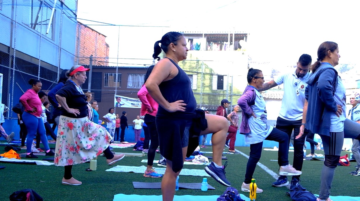 A imagem mostra um grupo de mulheres participando de uma atividade física ao ar livre, em uma quadra esportiva cercada por prédios residenciais.  Elas estão organizadas em fileiras, sobre tapetes ou toalhas no chão, realizando um exercício de equilíbrio, com uma perna apoiada no solo e a outra elevada. A maioria usa roupas confortáveis para prática esportiva, como leggings, camisetas e tênis. Algumas participantes vestem uniformes claros, possivelmente de um programa comunitário ou de saúde.  À direita, um profissional, provavelmente um educador físico ou agente de saúde, orienta os movimentos e auxilia uma das participantes. No fundo, outras pessoas observam ou também participam da atividade.  O ambiente sugere uma ação comunitária voltada à promoção da saúde, bem-estar e convivência, em um espaço público do bairro.