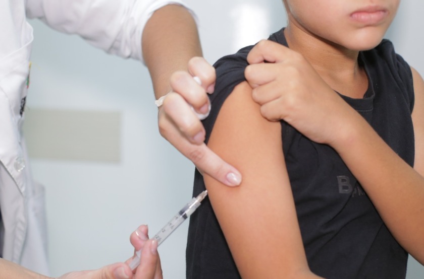 A imagem mostra uma criança sentada enquanto recebe uma vacina no braço. Um profissional de saúde, usando luvas e jaleco, aplica a injeção com uma seringa no braço da criança, que segura a própria camisa para facilitar o procedimento. O rosto da criança aparece parcialmente, com expressão séria, e o ambiente parece ser um local clínico, como um posto de saúde ou consultório.