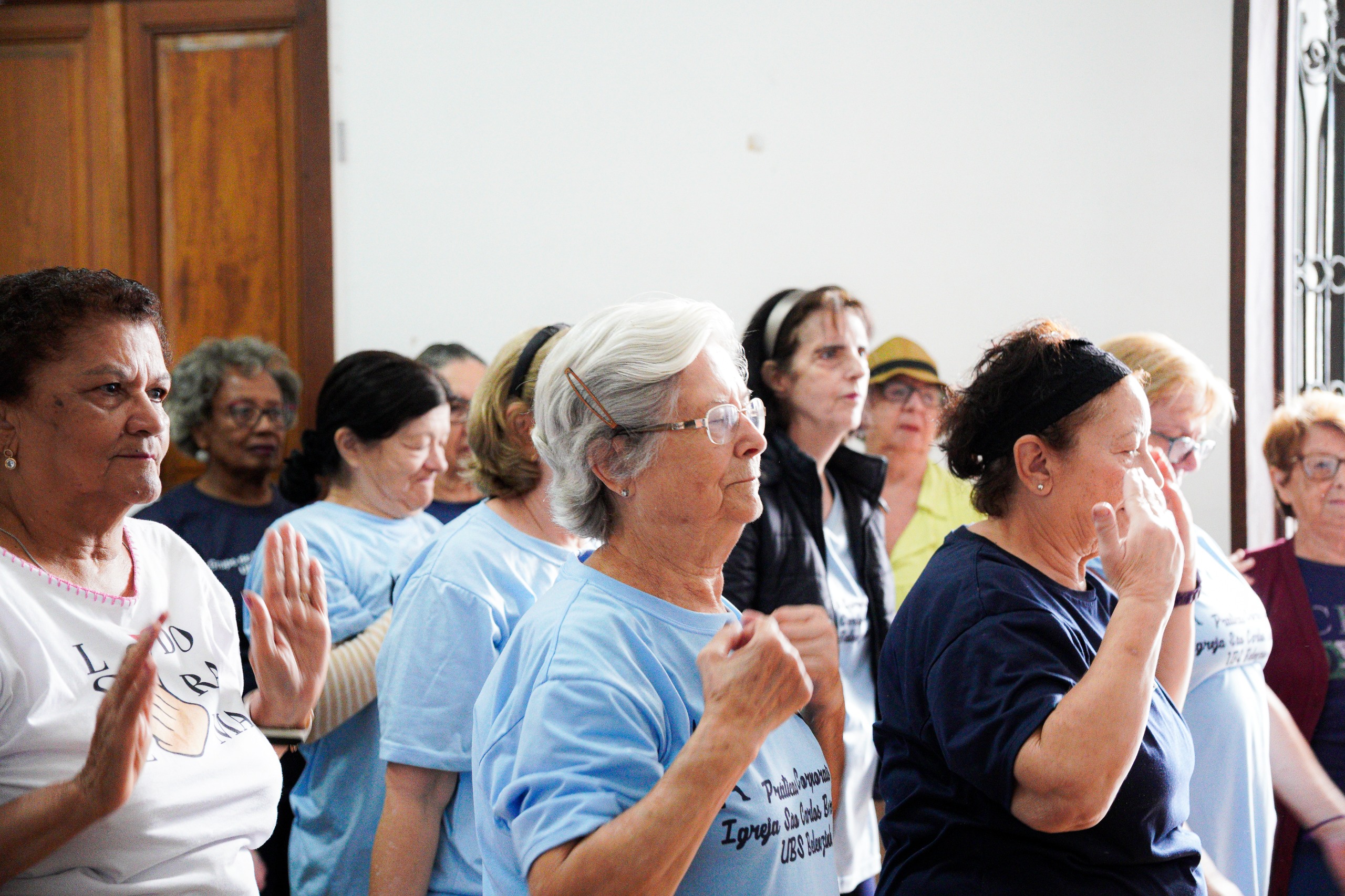 A cena retrata usuárias da UBS Belenzinho participando das atividades físicas da Academia da Saúde, iniciativa que promove práticas corporais, convivência e bem-estar. A atividade simboliza o fortalecimento das ações de promoção da saúde na cidade, em um momento em que o programa foi ampliado, passando de 18 para 66 polos na capital paulista. Elas estão lado a lado, realizando movimentos coordenados com os braços, aparentemente seguindo orientações de uma prática de exercícios ou alongamento.  Muitas usam camisetas claras — algumas em azul — possivelmente relacionadas à atividade ou ao grupo de saúde. As expressões são de concentração e engajamento, indicando atenção aos movimentos. Ao fundo, outras participantes acompanham o exercício, formando um grupo numeroso e diverso.