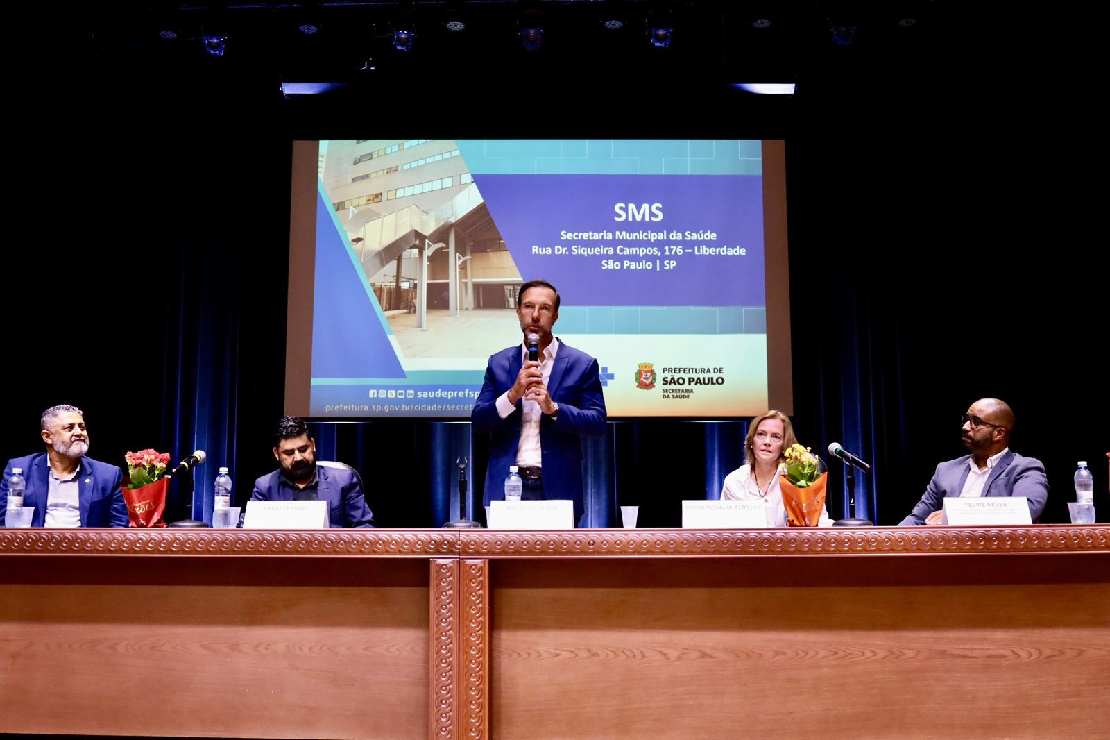 A imagem mostra uma mesa de evento ou cerimônia institucional em um auditório, com cinco pessoas posicionadas atrás de uma longa mesa de madeira sobre um palco.  No centro, um homem em pé, vestindo terno azul e camisa clara, fala ao microfone, aparentemente fazendo um discurso ou apresentação. À sua frente há uma plaquinha de identificação e uma garrafa de água.  Sentados dos dois lados estão quatro participantes, dois à esquerda e dois à direita, todos vestidos de forma formal. Sobre a mesa há microfones, copos descartáveis, garrafas de água e pequenos arranjos de flores.