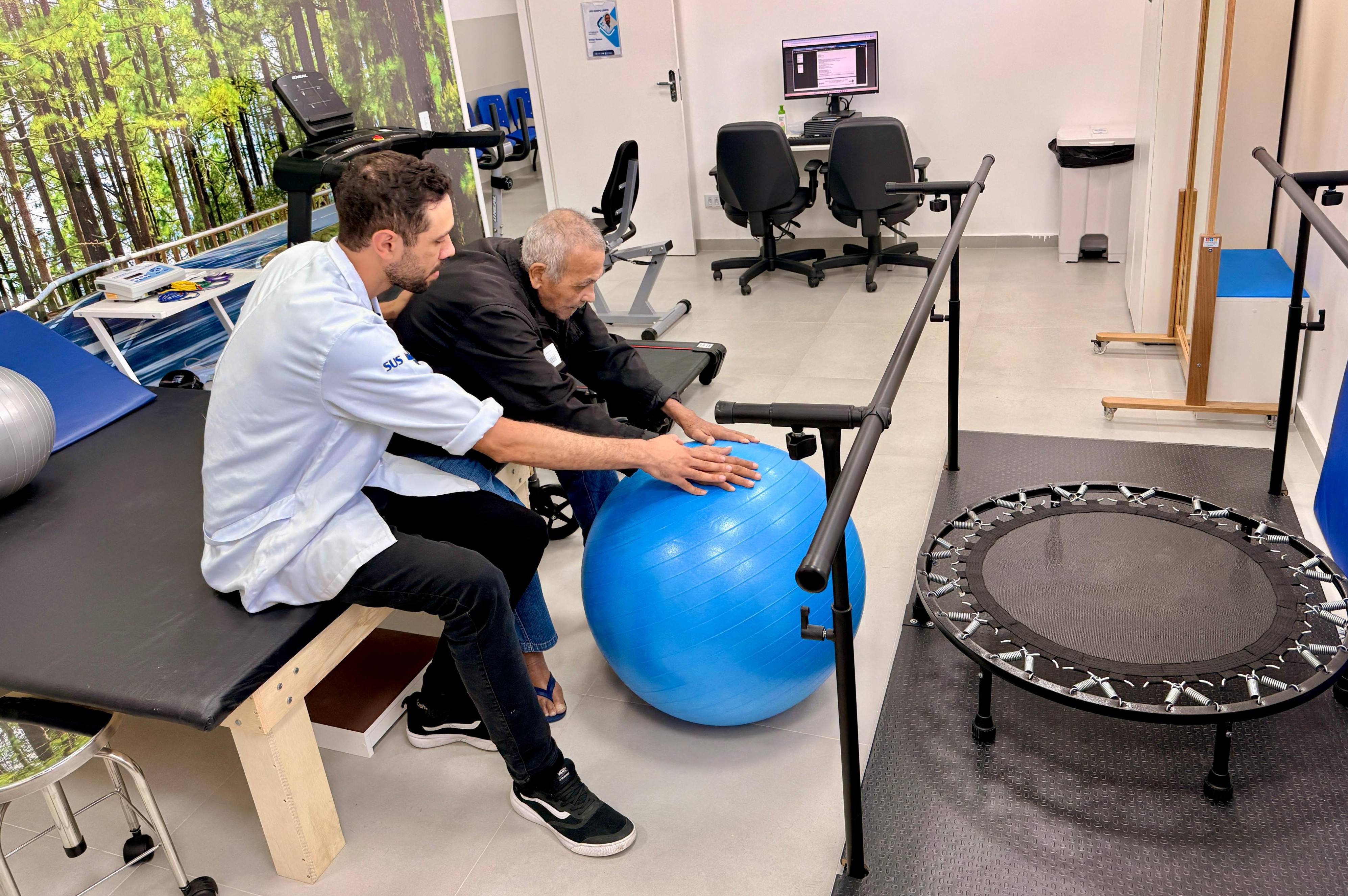 A imagem mostra uma sessão de fisioterapia realizada em um ambiente clínico de reabilitação. Em primeiro plano, um fisioterapeuta vestido com jaleco claro, com a sigla SUS no braço, acompanha um homem idoso sentado ao lado dele. O profissional orienta o paciente durante um exercício com uma bola terapêutica azul (bola suíça), apoiando as mãos sobre ela enquanto o idoso se inclina levemente para frente, indicando um exercício de fortalecimento ou equilíbrio. O espaço parece ser uma sala de fisioterapia ou reabilitação, equipada com diversos aparelhos. À direita, há barras paralelas para treino de marcha e um mini-trampolim de reabilitação. Ao fundo, é possível ver uma esteira ergométrica, equipamentos de exercício e um computador em uma mesa de atendimento com duas cadeiras. O ambiente é claro, organizado e bem iluminado, com paredes brancas e uma grande imagem decorativa de uma paisagem de floresta ao fundo, o que ajuda a tornar o espaço mais acolhedor.