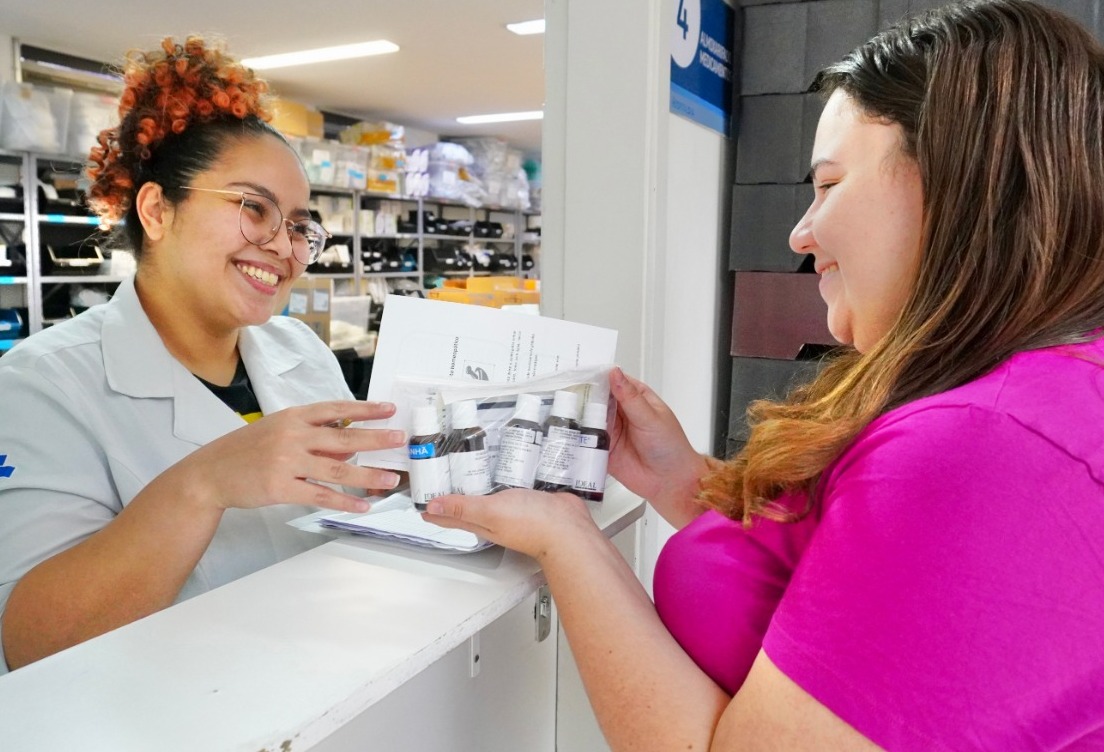 A imagem captura um momento de interação positiva em um ambiente de saúde ou farmácia. A foto mostra duas mulheres em um balcão de atendimento. A iluminação é clara e direta, típica de ambientes profissionais ou clínicos. Ao fundo, é possível ver estantes organizadas com diversas caixas e suprimentos, sugerindo que o local seja uma farmácia hospitalar, um almoxarifado de medicamentos ou uma unidade de saúde
