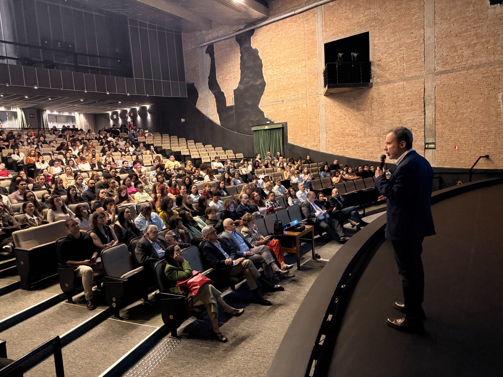 A imagem mostra um auditório amplo e cheio, durante uma apresentação ou palestra. No primeiro plano, à direita, um homem de terno escuro está de pé no palco segurando um microfone, aparentemente falando ao público. Ele está de perfil, voltado para a plateia, gesticulando enquanto se dirige às pessoas presentes. O auditório está praticamente lotado, com dezenas de pessoas sentadas em fileiras inclinadas de poltronas. O público é diverso em idade e aparência, e muitos parecem atentos ao que está sendo dito, olhando para o palestrante. Na primeira fileira, algumas autoridades ou convidados parecem estar sentados, vestidos de forma mais formal. Sobre uma pequena mesa à frente deles há um projetor, sugerindo que a apresentação pode incluir slides. A arquitetura do espaço chama atenção: as paredes laterais são de tijolo aparente, e há grandes elementos artísticos escuros aplicados na parede, que lembram silhuetas humanas estilizadas. A iluminação é suave e focada no palco e na plateia, típica de um teatro ou auditório institucional.