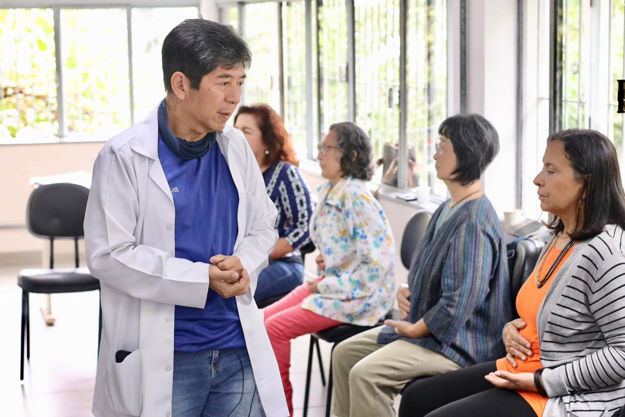 A foto mostra um ambiente interno iluminado por janelas amplas ao fundo. Em primeiro plano, à esquerda, um homem de traços asiáticos, cabelos curtos e escuros, veste um jaleco branco sobre uma camiseta azul e calça jeans. Ele está em pé, com as mãos unidas à frente do corpo, em uma postura de quem explica algo com atenção. À direita, quatro mulheres estão sentadas em cadeiras individuais, formando uma fila ou círculo de conversa. Elas parecem praticar um exercício de respiração consciente ou meditação, com as mãos repousadas sobre o abdômen e olhos fechados ou semicerrados. O clima é de tranquilidade, foco e aprendizado coletivo.