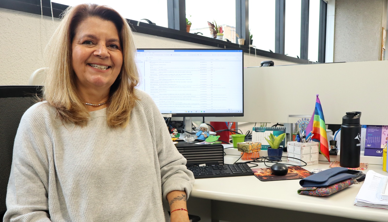 A imagem apresenta Rosane Jacy Fretes Fava em seu ambiente de trabalho. Ela é uma mulher de pele clara, com cabelos loiros de comprimento médio e um sorriso acolhedor. Veste uma blusa de tricô cinza claro. Rosane está sentada em sua mesa de escritório; ao fundo, vê-se um monitor de computador com planilhas, uma pequena bandeira do arco-íris (símbolo LGBTQIA+), itens de papelaria e uma planta pequena. Ela possui uma tatuagem delicada no antebraço esquerdo e usa uma pulseira de fio vermelho no pulso. A iluminação é natural, vinda de janelas amplas ao fundo.