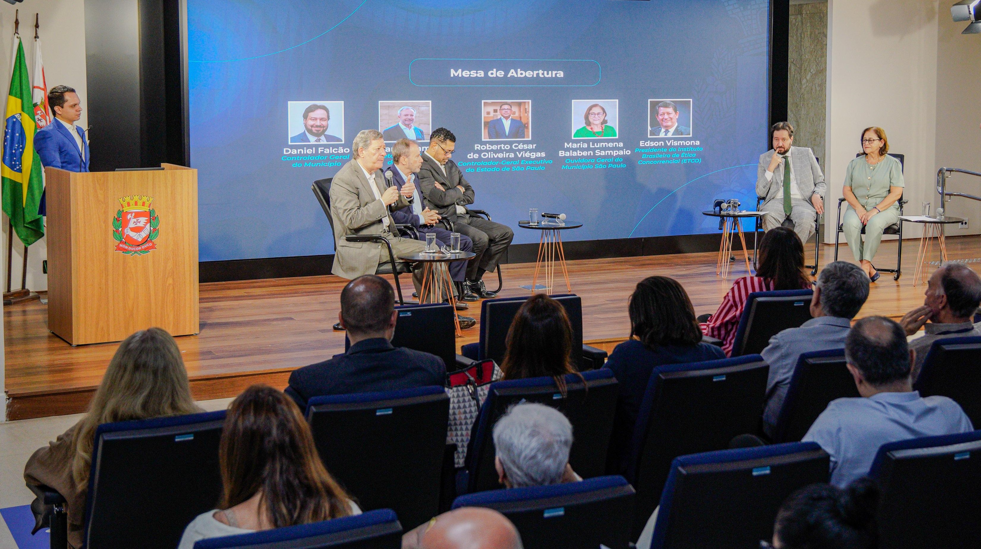 A imagem mostra um evento institucional realizado em um auditório, com público presente e uma mesa de abertura composta por autoridades. No palco, há cinco participantes sentados em cadeiras, enquanto um homem fala ao microfone em um púlpito ao lado esquerdo, próximo às bandeiras do Brasil e do estado/município.  Ao fundo, um grande telão exibe o título “Mesa de Abertura” e fotos com nomes e cargos dos participantes, indicando que se trata de um encontro formal ligado à gestão pública, possivelmente nas áreas de controle, ouvidoria ou administração. Os integrantes da mesa estão em traje social e participam de um debate ou apresentação, com microfones e mesas de apoio à frente.