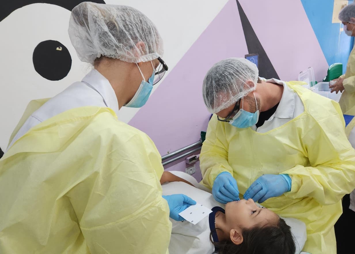 O Dia B da Saúde Bucal”, realizado na última sexta-feira (20) pelas equipes de Saúde Bucal da Secretaria Municipal da Saúde (SMS) em seis escolas da rede municipal de ensino, realizou um total de 728 atendimentos a crianças e adolescentes, entre escovação supervisionada, aplicação tópica de flúor, verniz de flúor, selante e tratamento restaurador atraumático (ART). Marta Cipriano, coordenadora de Saúde Bucal da SMS, explica que as intervenções foram realizadas de acordo com a triagem de risco odontológica previamente realizada como parte do Programa Saúde na Escola (PSE), parceria da SMS com a Secretaria Municipal da Educação (SME). “Estudantes considerados de baixo risco, sem problemas de saúde bucal detectados, receberam orientações de higiene oral e aprenderam a escovação correta; para os alunos de médio e alto risco, fizemos aplicação de flúor, de cariostático e o tratamento restaurador atraumático (ART), um procedimento odontológico não invasivo indicado para paralisar cáries, em que o dentista remove tecido cariado apenas com instrumentos manuais, selando a cavidade com cimento de ionômero de vidro”, explica a gestora.  Na cidade de São Paulo, o índice de cárie entre crianças de 5 anos de idade é de 0,97, e 0,49 na faixa de de 12 anos de idade. Em 2025, um total de 362.448 alunos de Escolas Municipais de Educação Infantil (EMEIs) e Escolas Municipais de Educação Fundamental (EMEFs) passaram por triagem odontológica dentro do PSE, que realizou 577.858 ações em Saúde Bucal. Ao longo do Dia B, as 32 equipes de Saúde Bucal de 25 Unidades Básicas de Saúde (UBSs) da capital realizaram 356 ARTs em 192 crianças. A assessora técnica de Saúde Bucal Thais Staut Zukeran explica que a aplicação do ART no ambiente escolar contribui para a diminuição do absenteísmo e favorece a continuidade do cuidado. “A estratégia permite a intervenção precoce em lesões de cárie, evitando a evolução, ao mesmo tempo em que preserva a estrutura dentária; além disso, por não exigir anestesia e ocorrer em um ambiente familiar, tende a ser melhor aceita pelas crianças, reduzindo medo e ansiedade.