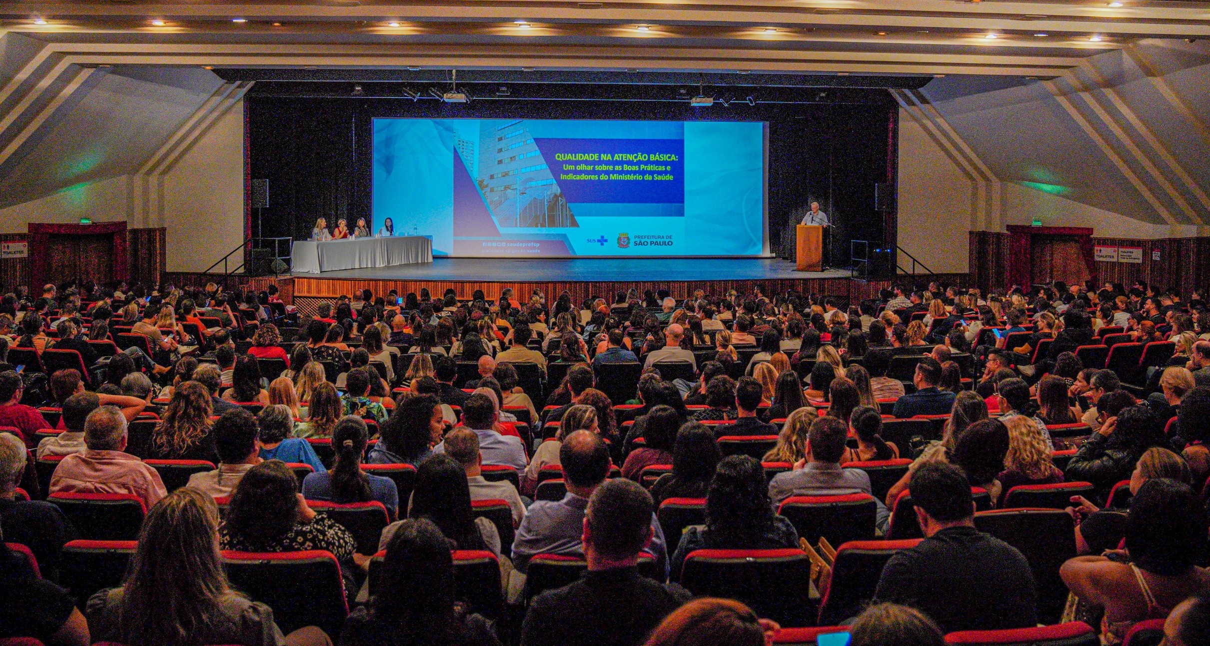 A imagem mostra um grande auditório lotado durante um evento institucional, com dezenas de pessoas sentadas em fileiras de poltronas vermelhas, todas voltadas para o palco. No palco, há uma mesa com quatro participantes sentados à esquerda e um palestrante em pé à direita, falando em um púlpito. Ao fundo, um grande telão exibe uma apresentação com o título “Qualidade na Atenção Básica: um olhar sobre as boas práticas e indicadores do Ministério da Saúde”, além da identidade visual da Prefeitura de São Paulo. O ambiente é bem iluminado e organizado, com estrutura de conferência, indicando um encontro técnico ou seminário voltado a profissionais da área da saúde, com foco em qualificação, indicadores e melhoria dos serviços públicos.
