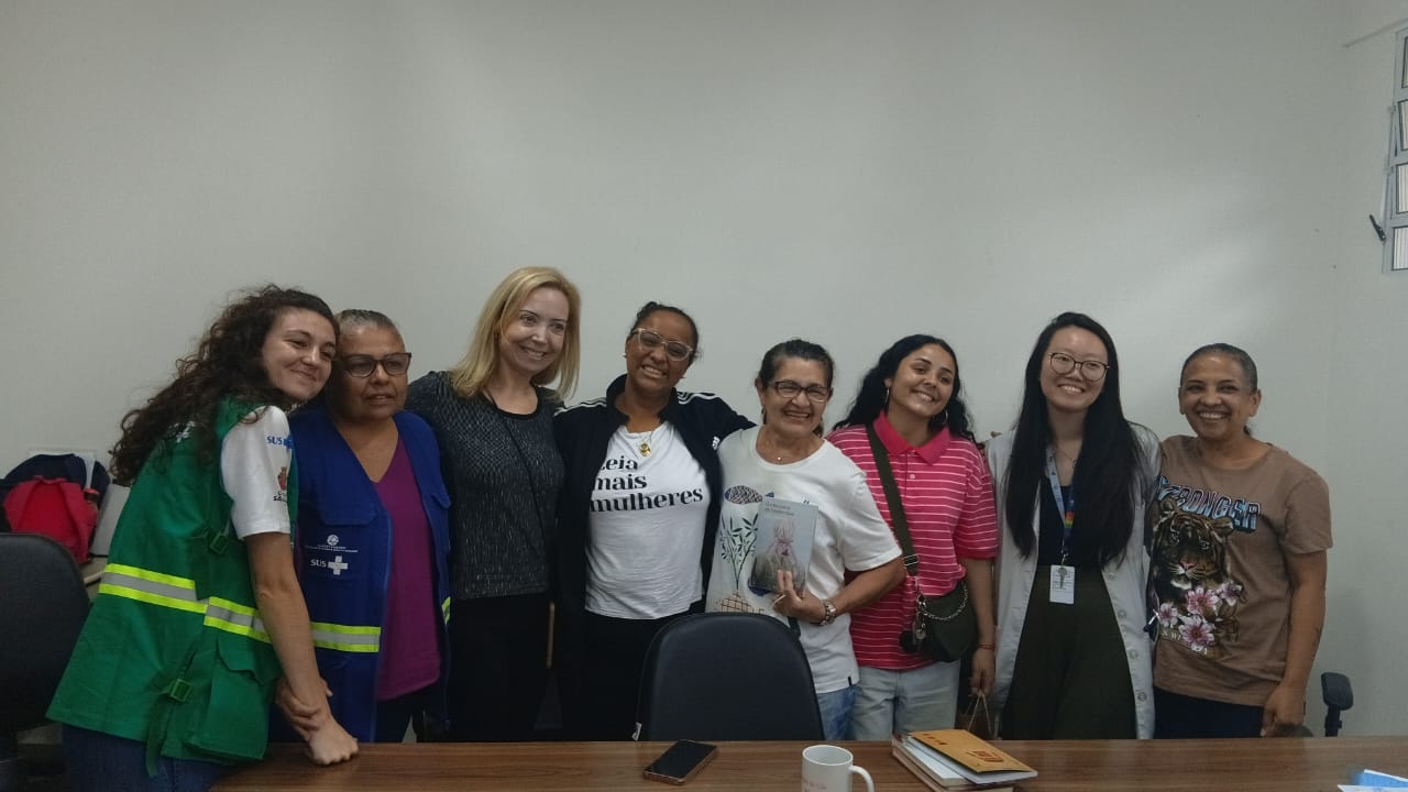 A imagem mostra um grupo de mulheres reunidas em uma sala simples, com parede branca e mesa de madeira à frente, dentro da UBS Parque Araribá. Ao centro está a escritora Lilia Guerra, que sorri enquanto posa junto às participantes. Ela está rodeada pelas idealizadoras e integrantes do clube de leitura da UBS Parque Araribá, formando uma composição afetiva e coletiva.  Algumas mulheres usam crachás e vestimentas que remetem ao trabalho na saúde, enquanto outras estão com roupas casuais. Uma delas segura um livro, reforçando o contexto literário do encontro. Sobre a mesa, há objetos como um celular, uma caneca e alguns livros empilhados, indicando um momento de troca e conversa.  A cena transmite proximidade, protagonismo feminino e valorização da leitura como prática de cuidado e convivência dentro do espaço da unidade de saúde.