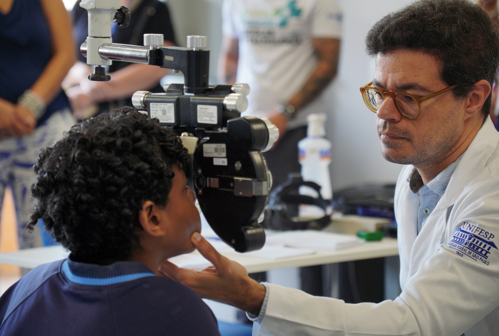 A imagem mostra um atendimento oftalmológico sendo realizado em uma criança. O paciente, um menino, está sentado diante de um equipamento de exame de visão (provavelmente um autorrefrator ou parte de um exame oftalmológico), enquanto um médico o observa atentamente.  O profissional de saúde, usando jaleco branco e óculos, posiciona delicadamente a cabeça da criança com a mão, garantindo o alinhamento correto para o exame. Ele demonstra concentração e cuidado durante o procedimento. No jaleco, é possível ver o logotipo de uma instituição de ensino/saúde.  Ao fundo, há outras pessoas desfocadas, sugerindo um ambiente clínico ou uma ação de atendimento coletivo, como mutirão ou triagem. Sobre a mesa, aparecem itens típicos de consultório, como frascos de álcool ou produtos de higienização.