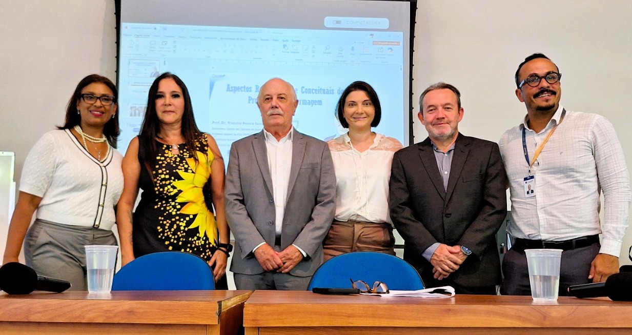 A imagem mostra seis pessoas em pé, lado a lado, posando para uma foto em um ambiente que parece ser uma sala de aula ou auditório.  Elas estão atrás de duas mesas de madeira com cadeiras azuis, onde há alguns objetos como copos de plástico, um microfone e óculos. Ao fundo, há um projetor exibindo um slide de apresentação — aparentemente relacionado a um tema técnico ou acadêmico, possivelmente na área de enfermagem.  O grupo é composto por quatro mulheres e dois homens, todos vestidos de forma profissional ou semiformal. No centro, um homem mais velho, de terno cinza, ocupa posição de destaque. Os demais estão distribuídos ao redor, alguns sorrindo levemente, outros com expressão neutra.