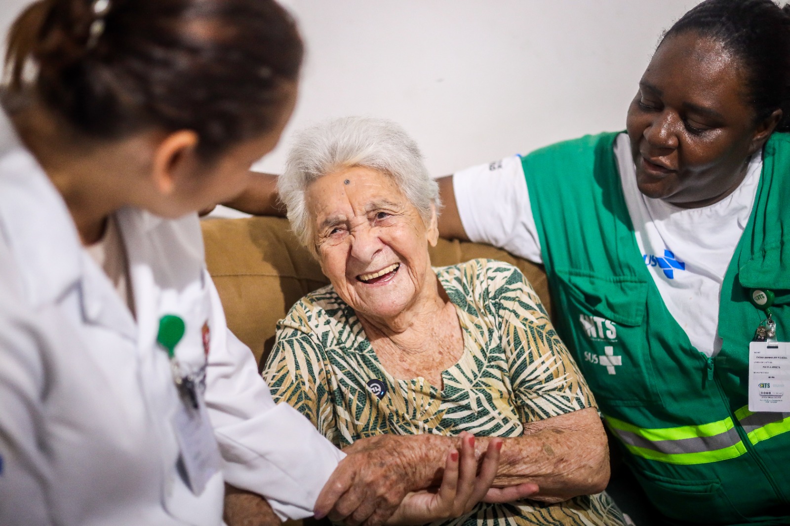 A fotografia mostra uma senhora idosa, Dona Carolina, sentada em um sofá, radiante com um sorriso largo e genuíno que ilumina seu rosto. Ela tem cabelos brancos curtos e veste uma blusa com estampa de folhagem em tons de verde e bege. Ao seu redor, duas profissionais de saúde realizam o atendimento: À esquerda: Uma profissional de jaleco branco segura gentilmente a mão de Dona Carolina, demonstrando uma conexão direta e afetuosa. À direita: Outra profissional, vestindo um colete verde com identificações que remetem ao sistema de saúde (SUS e NTS), observa a idosa com um olhar atento e acolhedor, mantendo o braço em volta do encosto do sofá, o que reforça a sensação de proteção. A iluminação é suave, focando na expressão de felicidade da paciente, criando uma atmosfera de confiança e dignidade.