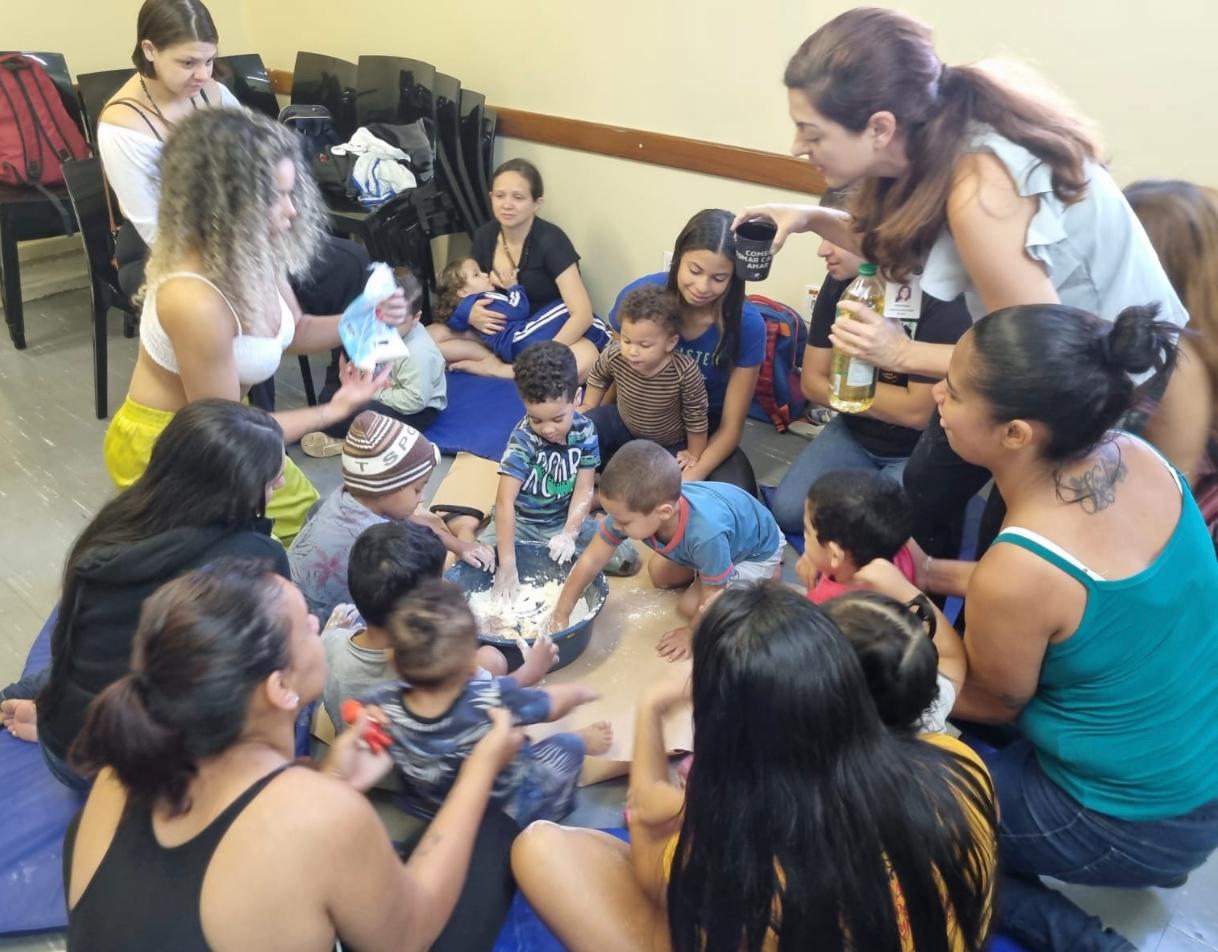 A imagem retrata uma cena vibrante e acolhedora de um grupo de estimulação infantil realizado na UBS Paraisópolis I. O foco central é a interação entre crianças pequenas, seus cuidadores e profissionais de saúde em uma atividade prática e sensorial.Um grupo de crianças e adultos está sentado em círculo sobre colchonetes azuis no chão. No centro, há uma bacia com farinha branca, onde as crianças estão colocando as mãos para explorar a textura. Uma profissional de saúde (à direita) adiciona óleo à mistura, indicando a preparação de uma 