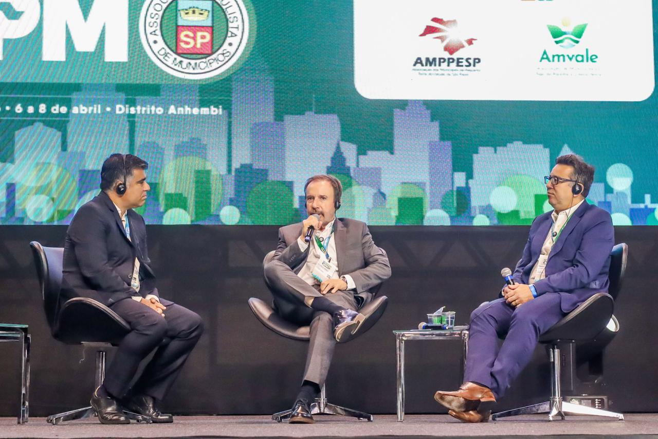 A imagem mostra um painel de debate em um evento institucional realizado em um auditório. Três homens estão sentados em poltronas no palco, todos usando fones de ouvido e crachás de identificação. O homem ao centro segura um microfone e parece estar falando, enquanto os outros dois o escutam atentamente.  Ao fundo, há um grande telão com a identidade visual do evento, em tons de verde e azul, com ilustrações de prédios ao estilo skyline. Também aparecem logotipos de entidades como a Associação Paulista de Municípios, a Associação dos Municípios do Vale do Paraíba e Litoral Norte e a Associação dos Municípios e Prefeitos do Estado de São Paulo. Na parte superior esquerda, é possível ler a referência ao período “6 a 8 de abril” e ao local “Distrito Anhembi”, indicando que se trata de um encontro ou congresso voltado à gestão pública ou municipal.