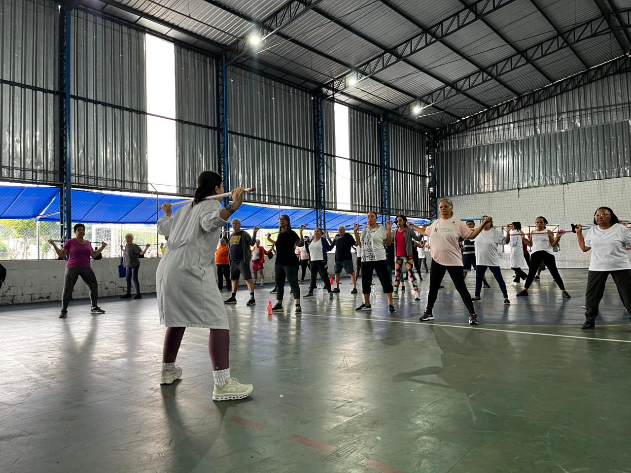 A foto registra uma aula de ginástica terapêutica em uma quadra poliesportiva coberta. Em primeiro plano, de costas para a câmera, uma profissional de saúde (possivelmente uma fisioterapeuta ou educadora física) veste um jaleco branco e guia o grupo. Ela segura um bastão sobre os ombros, demonstrando um exercício de postura e alongamento.  Ao fundo, cerca de 15 a 20 pacientes — em sua maioria idosos — seguem atentamente os movimentos. Eles estão espalhados pela quadra, mantendo distanciamento, e utilizam bastões coloridos para auxiliar nos exercícios. O ambiente é amplo, com iluminação natural vindo das laterais e teto alto de estrutura metálica, transmitindo uma sensação de vitalidade e cuidado comunitário.