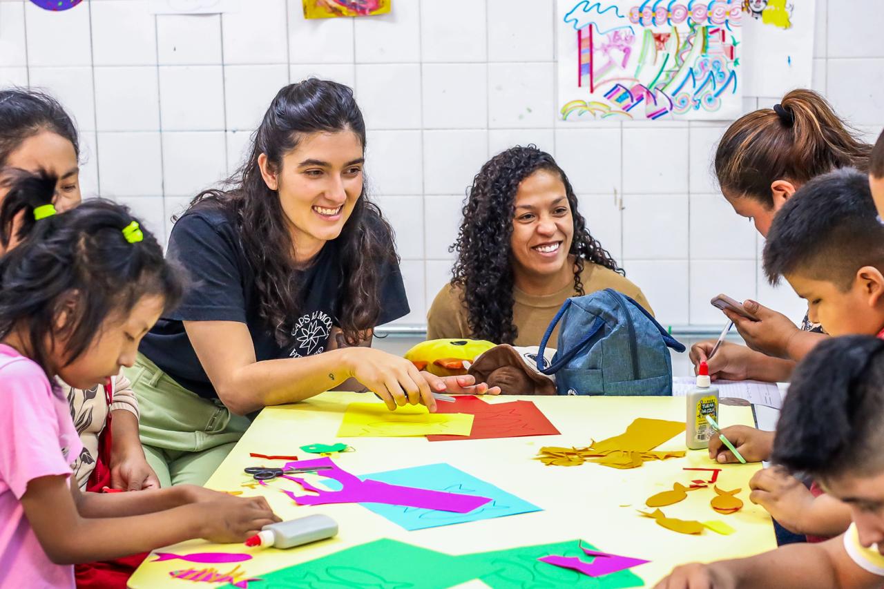 Grupo de crianças participa de uma atividade coletiva ao redor de uma mesa, acompanhadas por duas profissionais. As profissionais interagem com os participantes, orientando e incentivando a produção de desenhos e colagens com papéis coloridos. O ambiente é descontraído e colaborativo, com sorrisos e trocas entre crianças e adultas, reforçando a convivência, a integração e o cuidado em grupo.