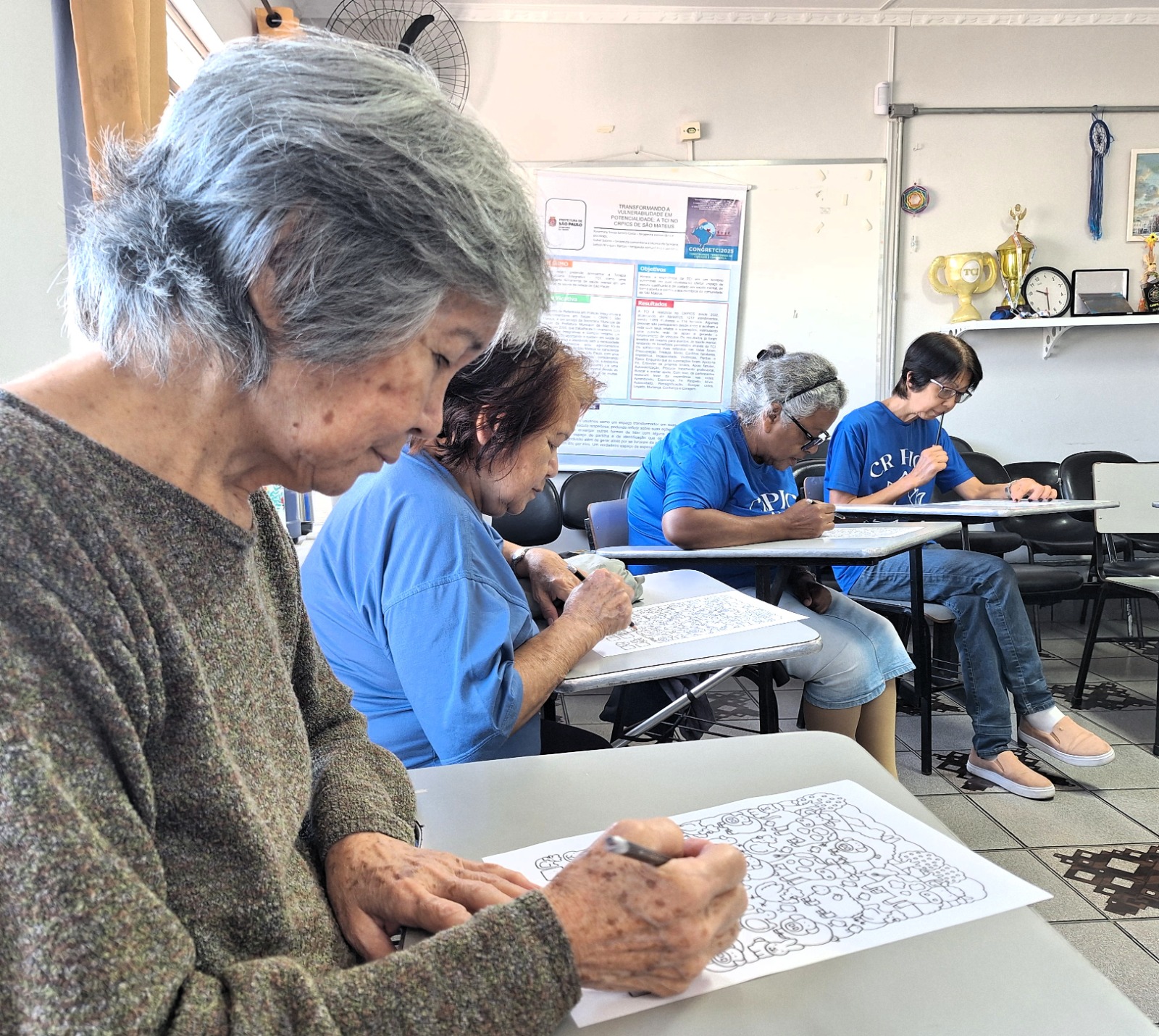 Em uma sala de atividades, quatro pessoas idosas participam de uma dinâmica da Academia da Memória, sentadas em carteiras escolares e concentradas em exercícios impressos. Em primeiro plano, uma mulher de cabelos grisalhos escreve atentamente em uma folha com desenhos e atividades cognitivas. Ao fundo, outras participantes também realizam as tarefas com foco, em um ambiente acolhedor e iluminado. Na parede, há cartazes informativos e prateleiras com troféus, sugerindo um espaço de convivência e aprendizagem.