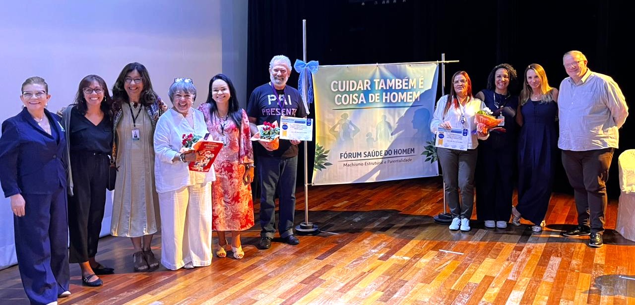 A imagem mostra um grupo de cerca de dez pessoas posando lado a lado em um palco, em um ambiente que parece ser um auditório ou teatro. O chão é de madeira e há iluminação direcionada, típica de eventos ou apresentações.  Ao centro, há um banner com a frase “Cuidar também é coisa de homem”, acompanhado do subtítulo “Fórum Saúde do Homem” e a menção a temas como machismo estrutural e parentalidade. O banner também traz silhuetas de figuras humanas, reforçando o tema do cuidado e da participação masculina na saúde.  As pessoas estão sorrindo, algumas com os braços apoiados umas nas outras, transmitindo um clima de celebração e reconhecimento. Algumas seguram certificados e buquês de flores, indicando que pode se tratar de uma homenagem, premiação ou encerramento de evento.