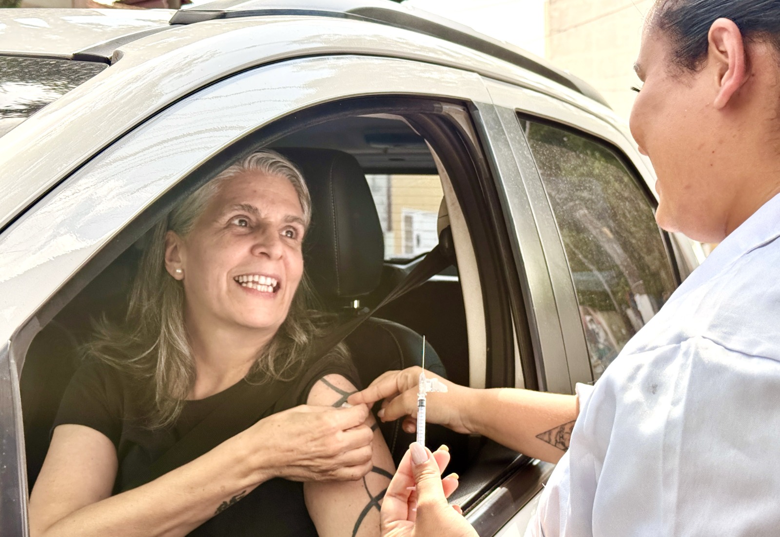 A imagem mostra uma mulher dentro de um carro, com o vidro da janela aberto, recebendo uma injeção no braço de uma profissional de saúde que está do lado de fora do veículo.  A mulher, de cabelos grisalhos e expressão sorridente, parece tranquila e à vontade, olhando para a profissional. Ela veste uma blusa preta e segura a manga para facilitar a aplicação. Já a profissional de saúde, usando uniforme claro, segura a seringa e aplica a vacina no braço da paciente.  A cena sugere um atendimento no modelo drive-thru de vacinação, em que a pessoa é imunizada sem precisar sair do carro. O ambiente parece ser ao ar livre, durante o dia, e transmite uma sensação de cuidado, acolhimento e eficiência no atendimento de saúde.