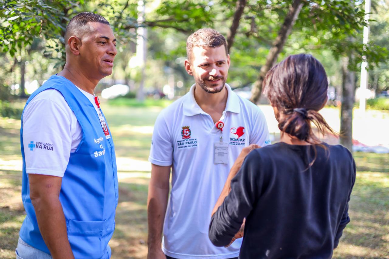 A imagem mostra três pessoas conversando em um parque arborizado durante o dia.  Dois profissionais de saúde estão em pé, voltados para uma mulher que aparece de costas para a câmera. À esquerda, um homem usa camiseta branca e colete azul com a identificação “Consultório na Rua”. No centro, outro profissional veste uma camisa polo branca com logotipos da Prefeitura de São Paulo e de um programa social, além de um crachá pendurado no pescoço. Ambos parecem atentos e receptivos durante a conversa.  A mulher, de cabelo preso e blusa preta, gesticula enquanto fala com os profissionais. O ambiente ao redor é iluminado por luz natural, com árvores e áreas verdes ao fundo, transmitindo um contexto de acolhimento e atendimento em espaço público.